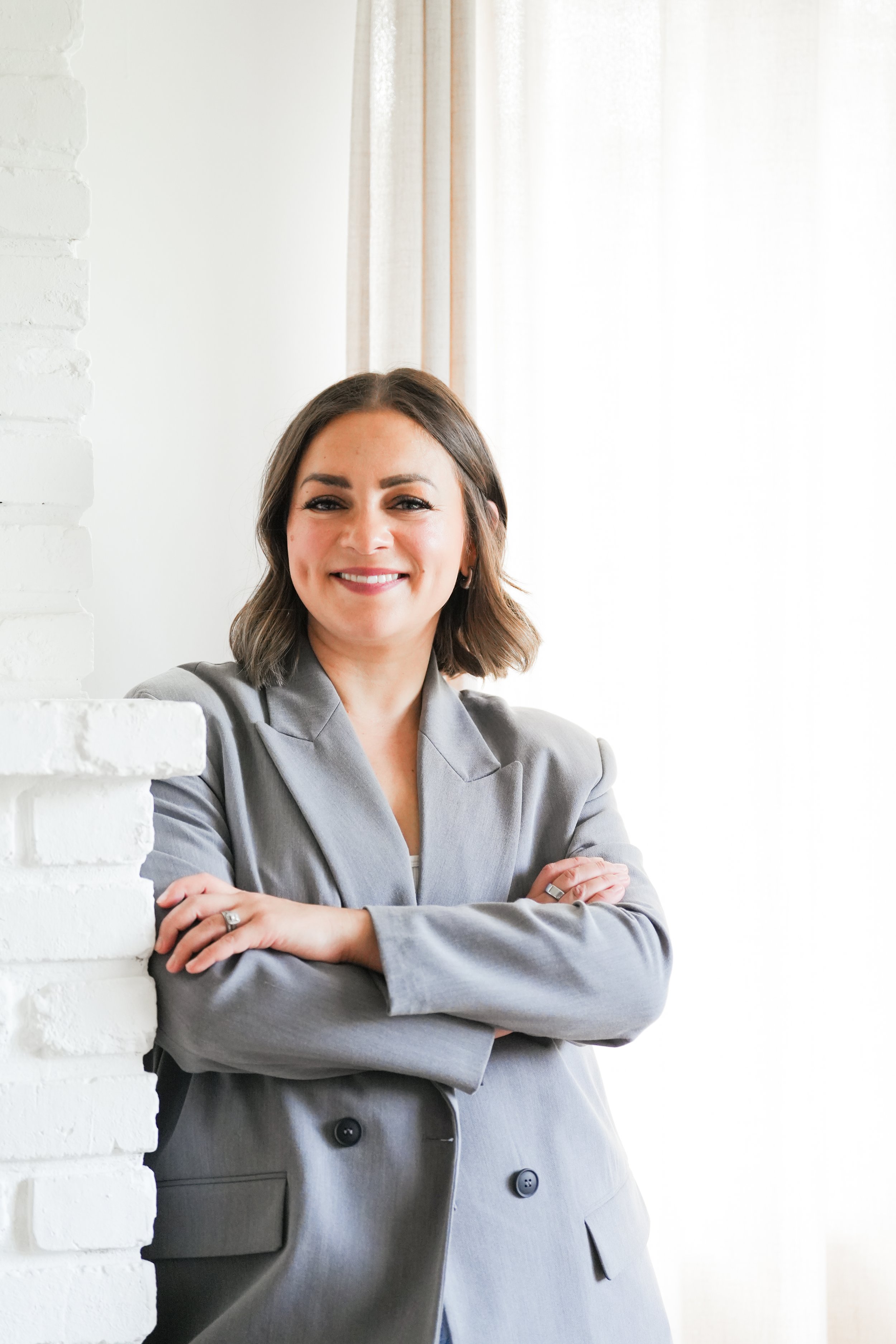 Bree Graham smiling with her arms crossed, wearing a gray blazer beside a window.