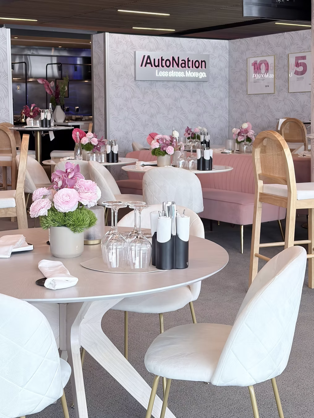 A stylish restaurant interior with round tables set with pink and white floral arrangements, wine glasses, and black and white cutlery holders. Pink cushioned benches and white upholstered chairs with gold legs provide seating. The decor features light-colored walls with framed posters and a sign for AutoNation.