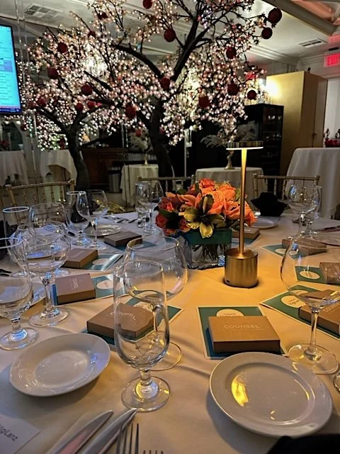 A decorated banquet table set with glassware, plates, and menus, arranged in front of a floral centerpiece with an ornate tree adorned with lights and red ornaments in the background.