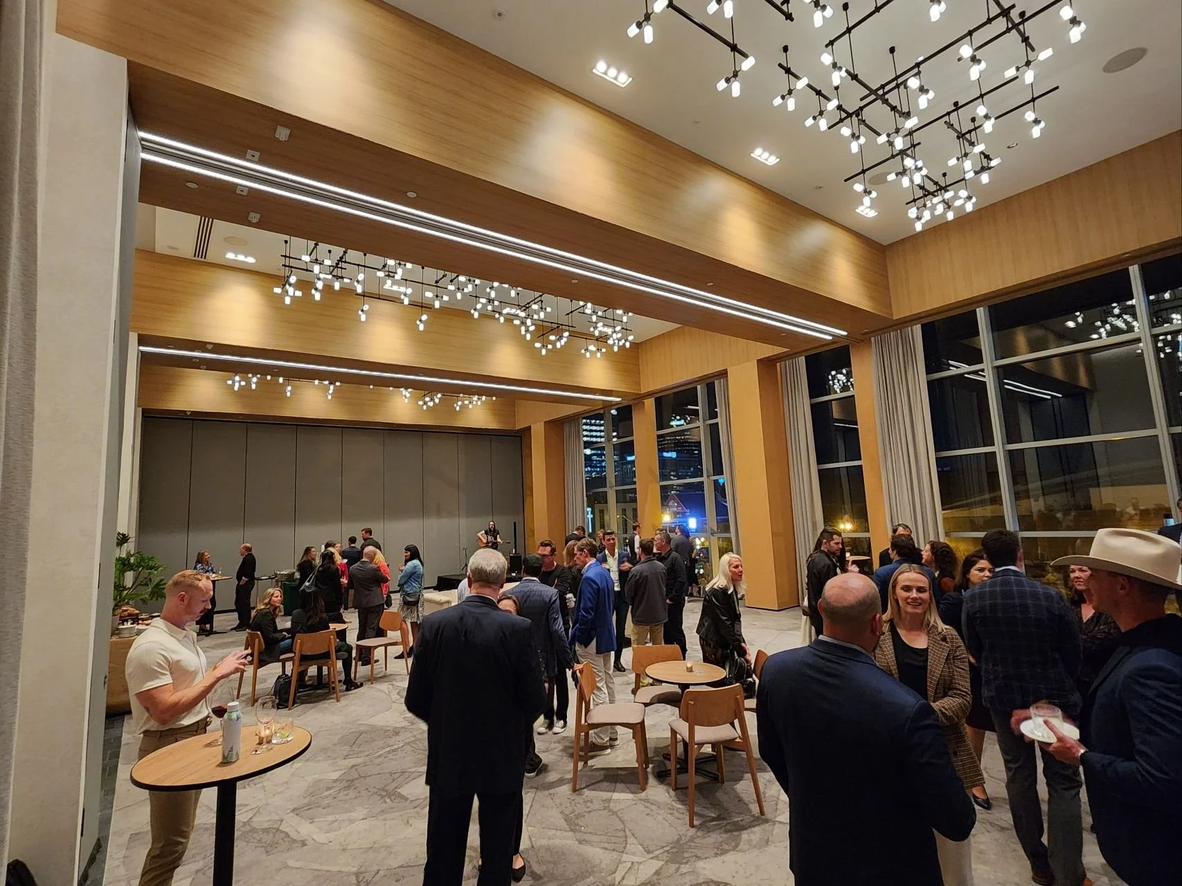 A large indoor event with many people socializing, some standing and others seated at tables. The room has high ceilings with modern patterned chandeliers, wooden accents, and floor-to-ceiling windows showing the night outside.