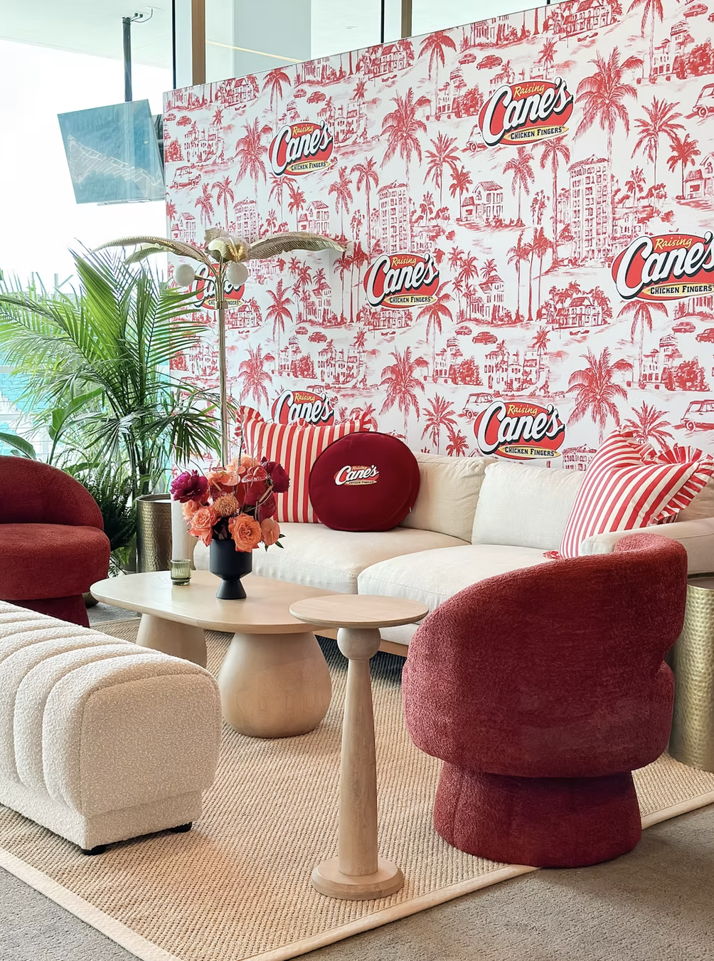 A stylish waiting area with a beige sofa adorned with red and white striped pillows, red velvet chairs, a white textured ottoman, a wooden coffee table with a floral arrangement, and a large green plant. The background features a Red and white patterned wall with the Raising Cane's logo and palm trees, suggesting a casual, modern restaurant or lounge setting.