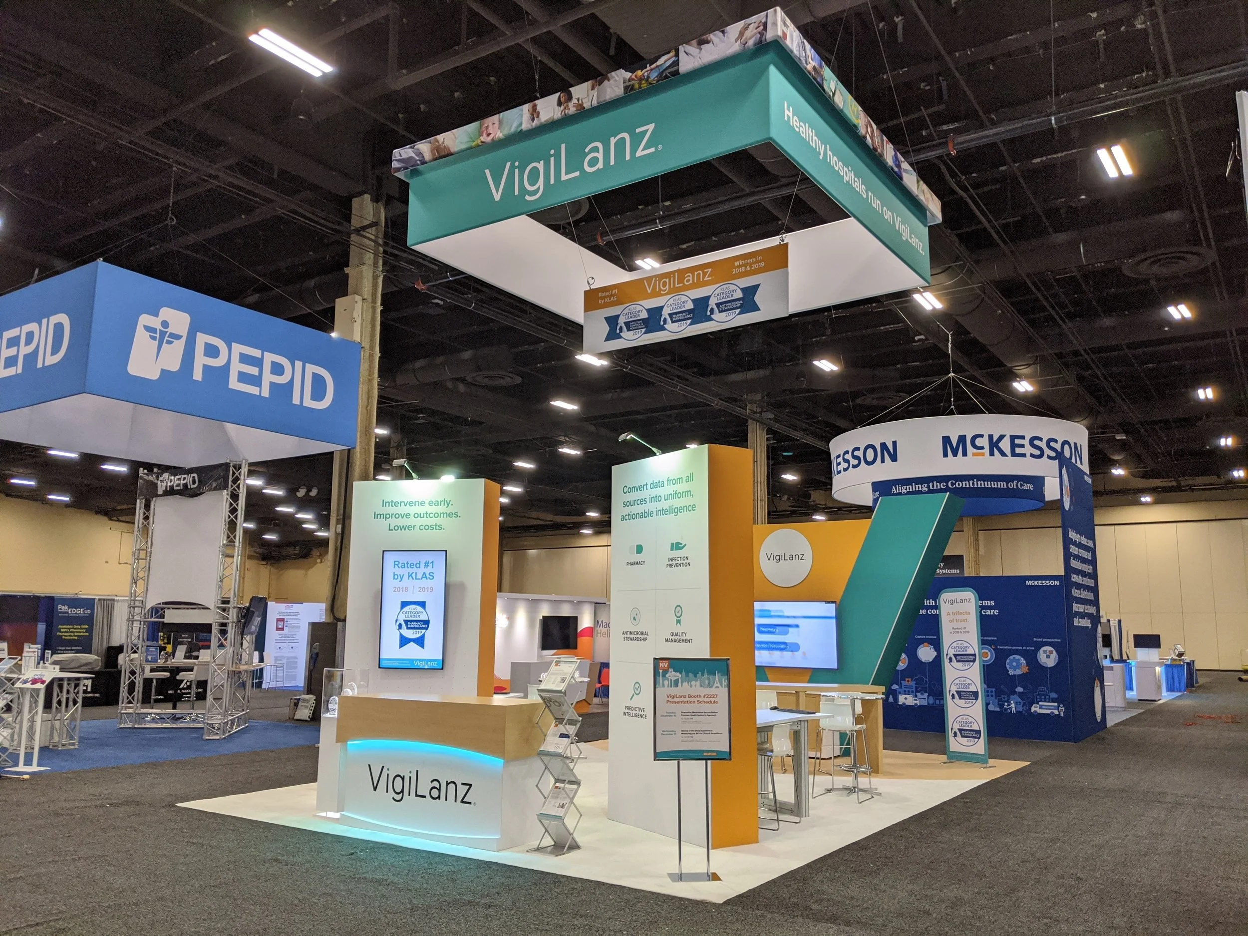An exhibition booth for Vigilanz at a trade show, featuring a teal and white color scheme with signage displaying 'Vigilanz' and 'Healthy hospitals run on Vigilanz.' Several other booths and displays are visible in the background within the large indoor convention center.