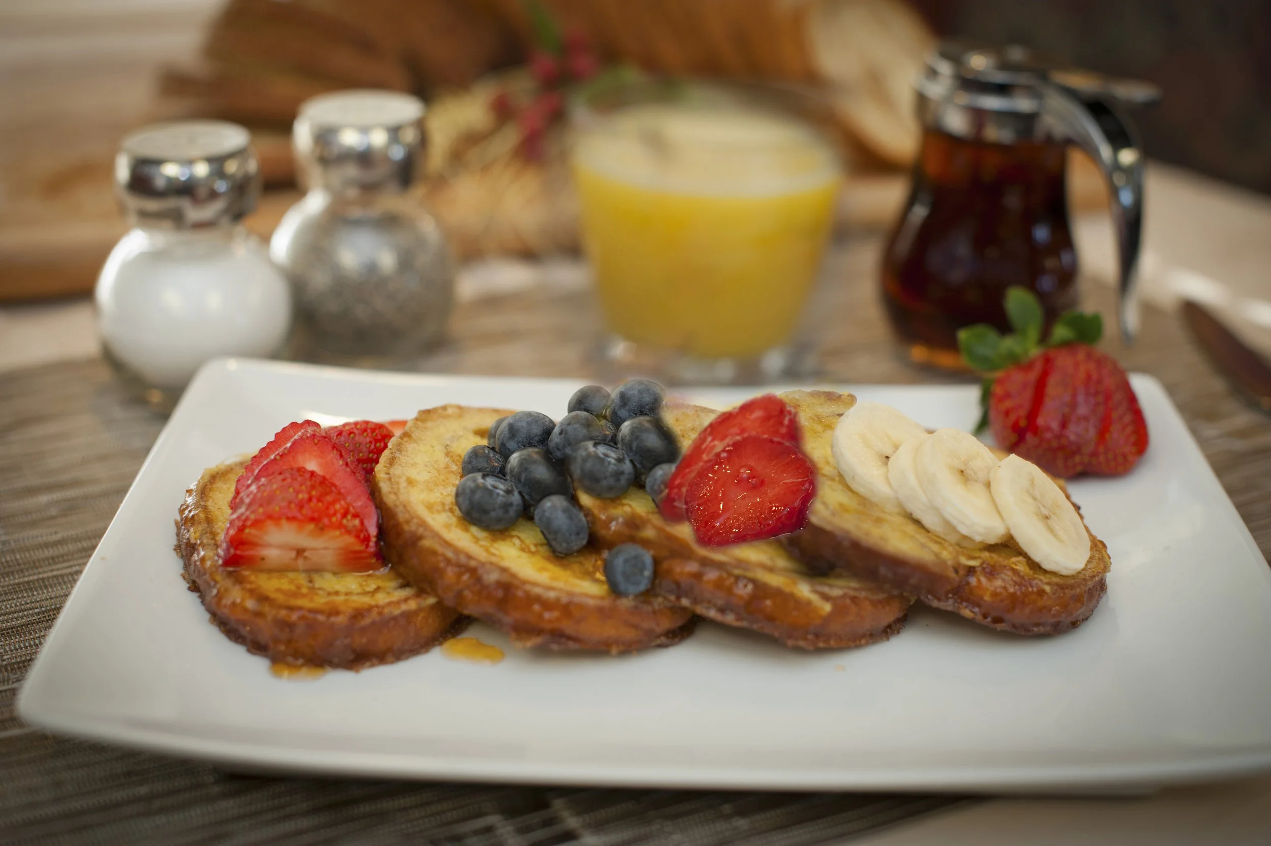 A close-up of a plate with French toast topped with strawberries, banana slices, and blueberries, set on a wooden table with a blurred background featuring condiments and a glass of orange juice.
