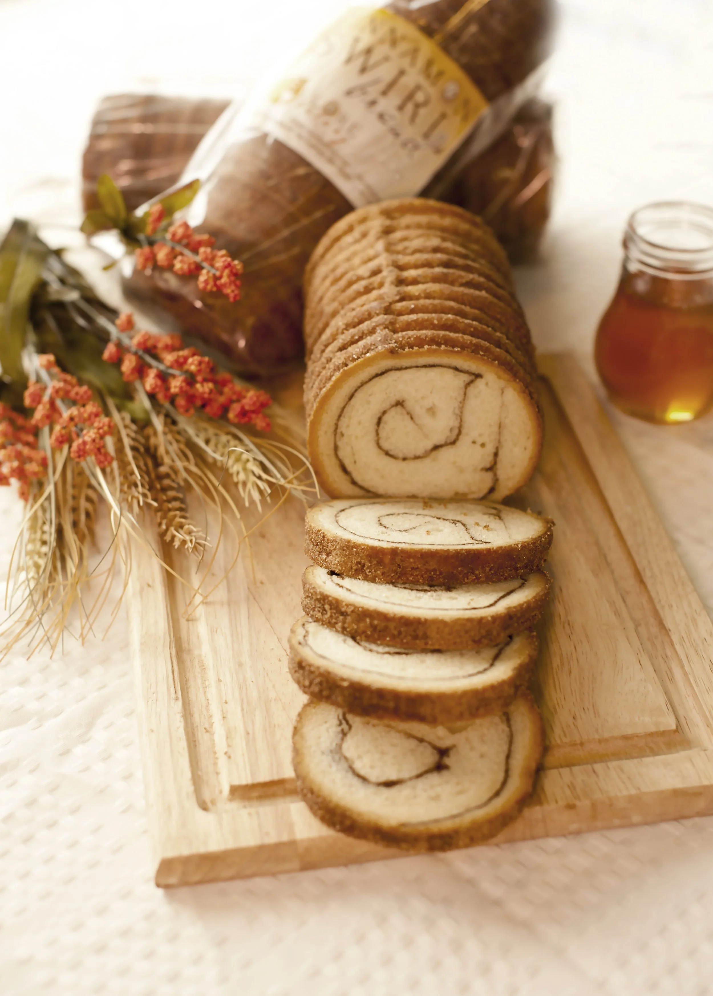 Sliced Swiss roll cake with a swirl pattern on a wooden serving board, decorated with wheat and orange flowers, and a small jar of honey in the background.