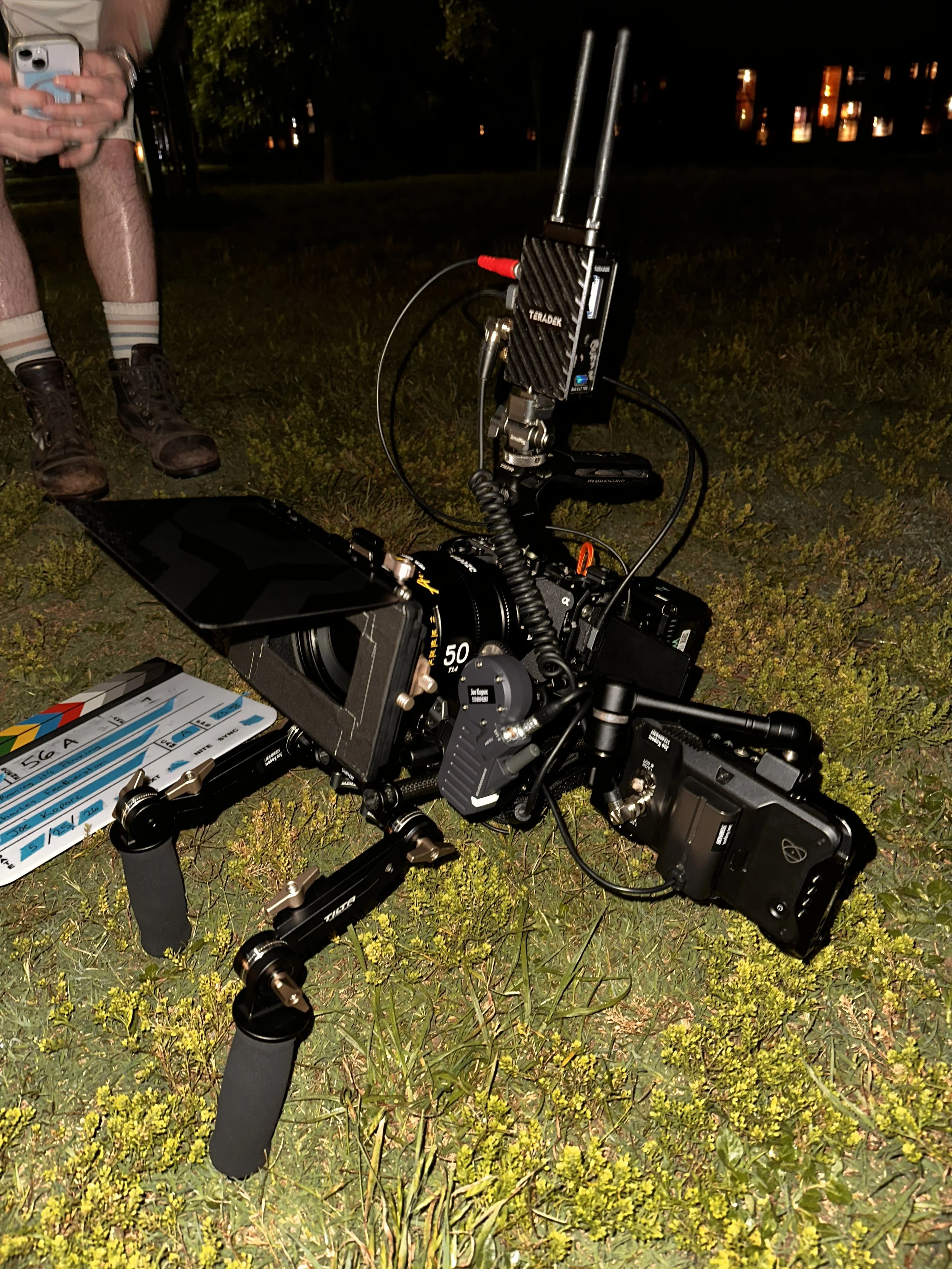 Nighttime outdoor scene with camera equipment and a person holding a smartphone, standing on grass with small yellow flowers, with a building lit in the background.