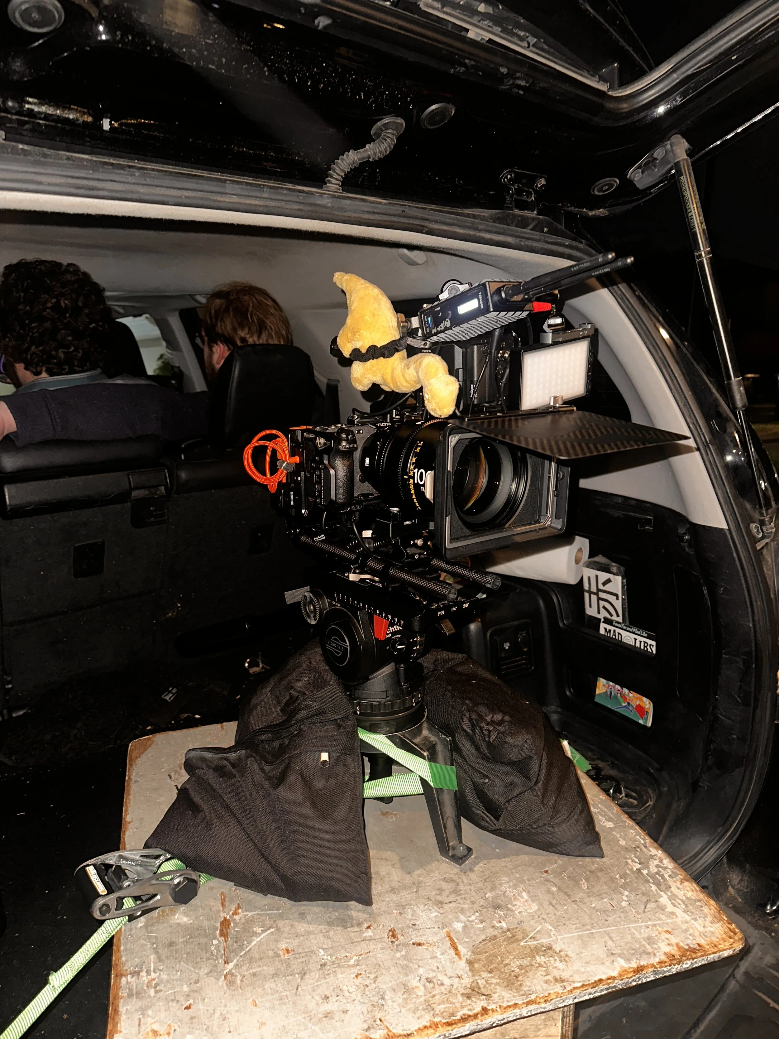 A professional video camera mounted on a tripod with a soft toy attached, set up inside a vehicle. The vehicle has a camera rig and two people are sitting in the front seats, possibly filming or preparing to film.
