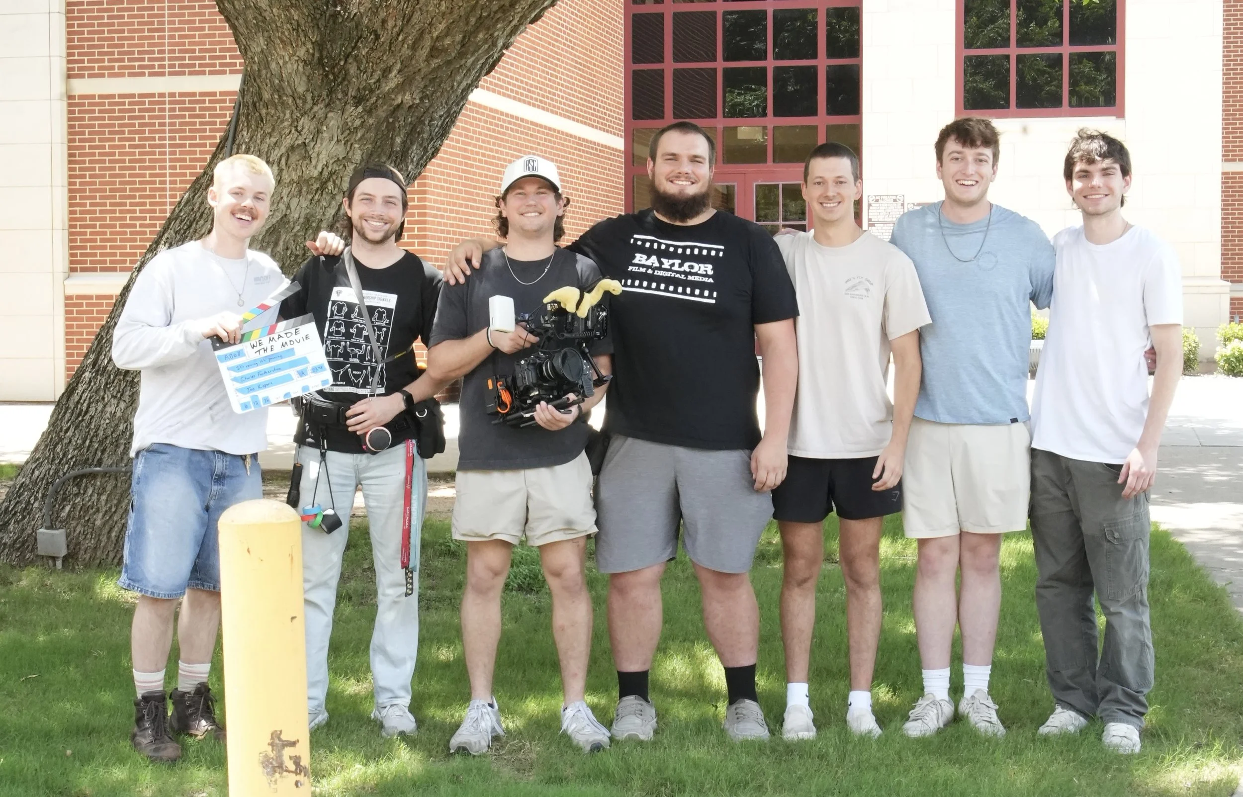 A group of seven young men stands outdoors in front of a large tree and a building with brick and white walls, smiling at the camera. They are casually dressed, and one holds a clapperboard, while another holds a professional video camera.