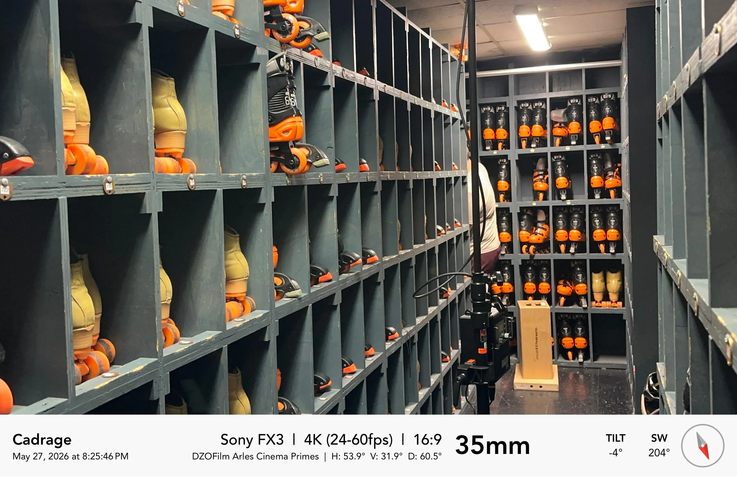 A storage room with multiple cubby shelves filled with inline skate shoes and orange skate wheel parts. There are some boxes and equipment, with a person partially visible in the background.