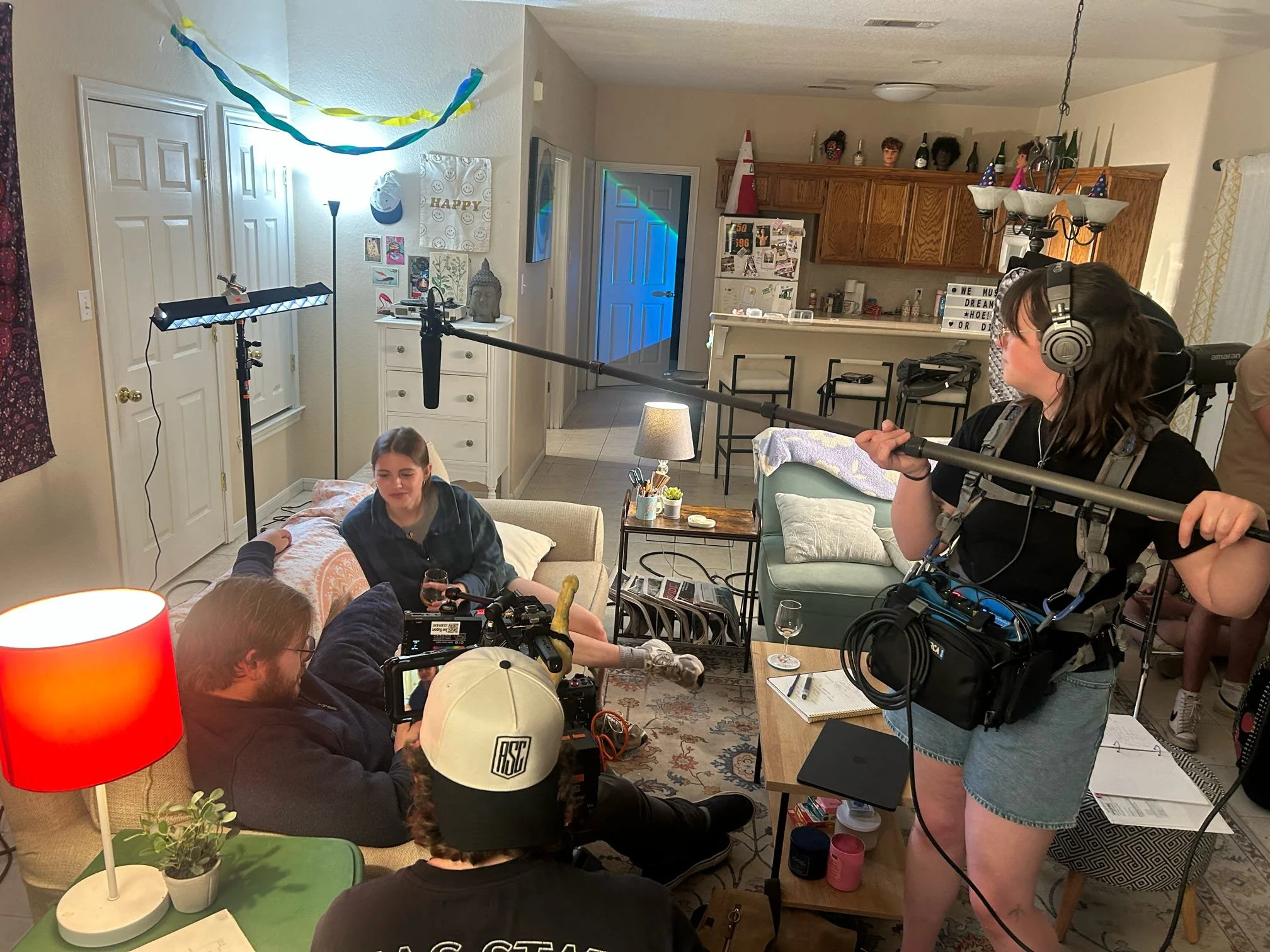 A group of people in a living room filming a video. There is a woman with headphones holding a boom microphone, a person with a camera, and multiple others either sitting or preparing to film. The room has decorations, furniture, and curtains, with a