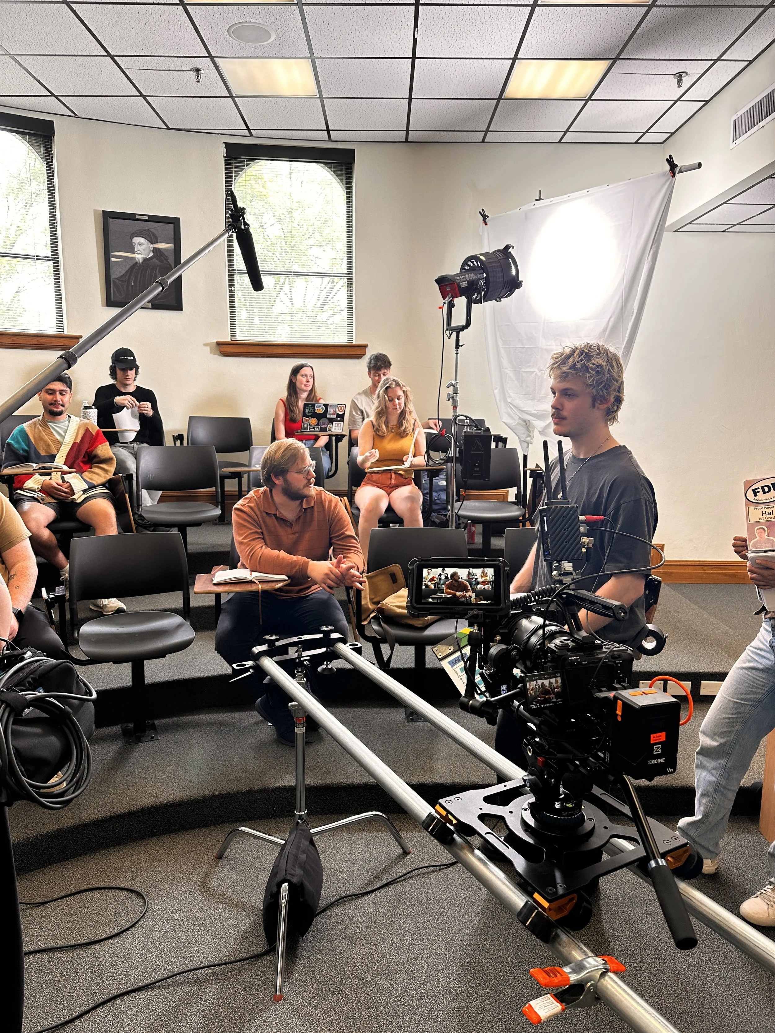 People in a meeting room with film equipment, including a camera on a dolly, lighting, and a white backdrop, actors and crew preparing for filming.