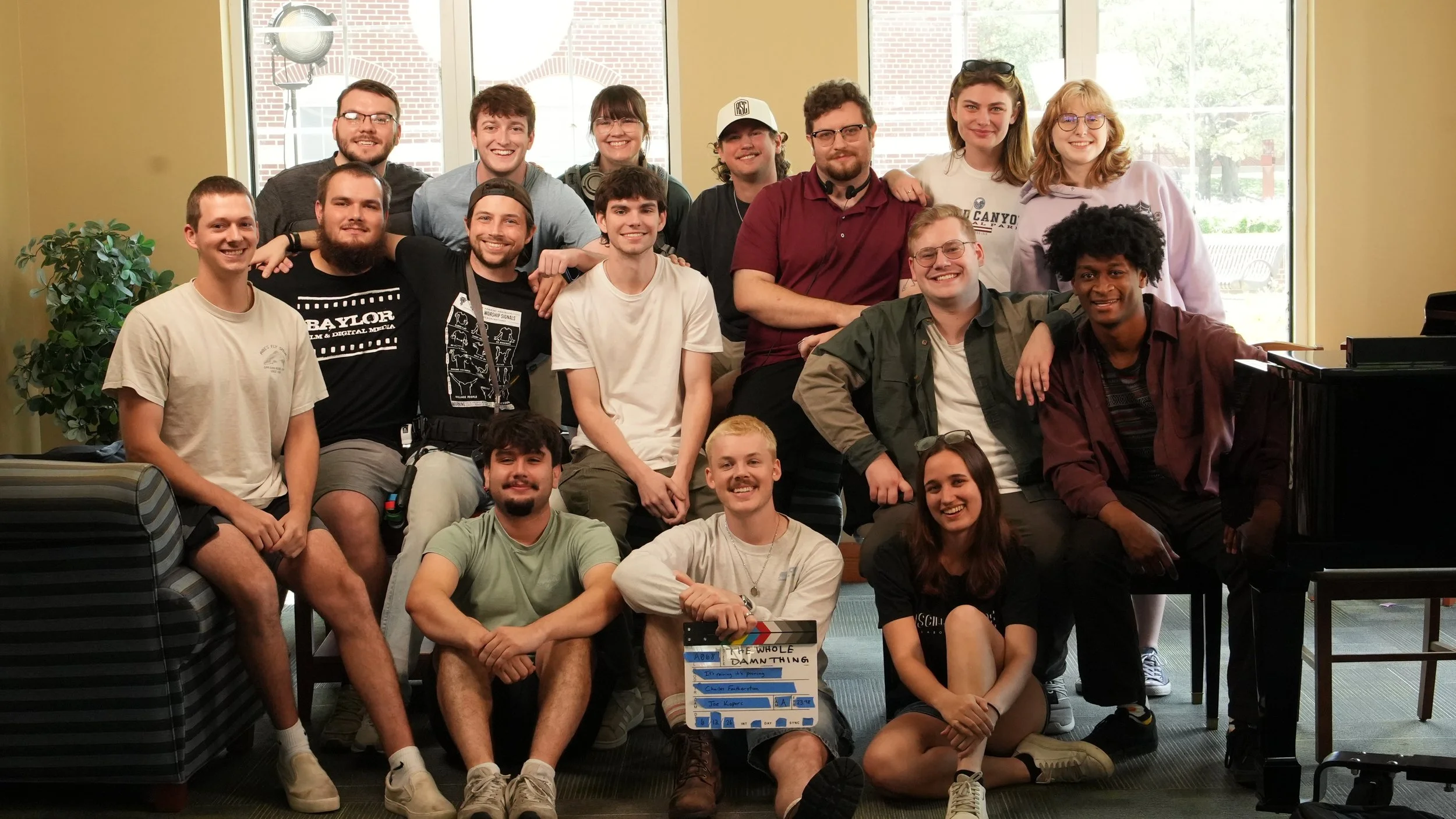 A group of young adults smiling and posing together in a room with large windows, some sitting and others standing, holding a clapperboard with writing on it.