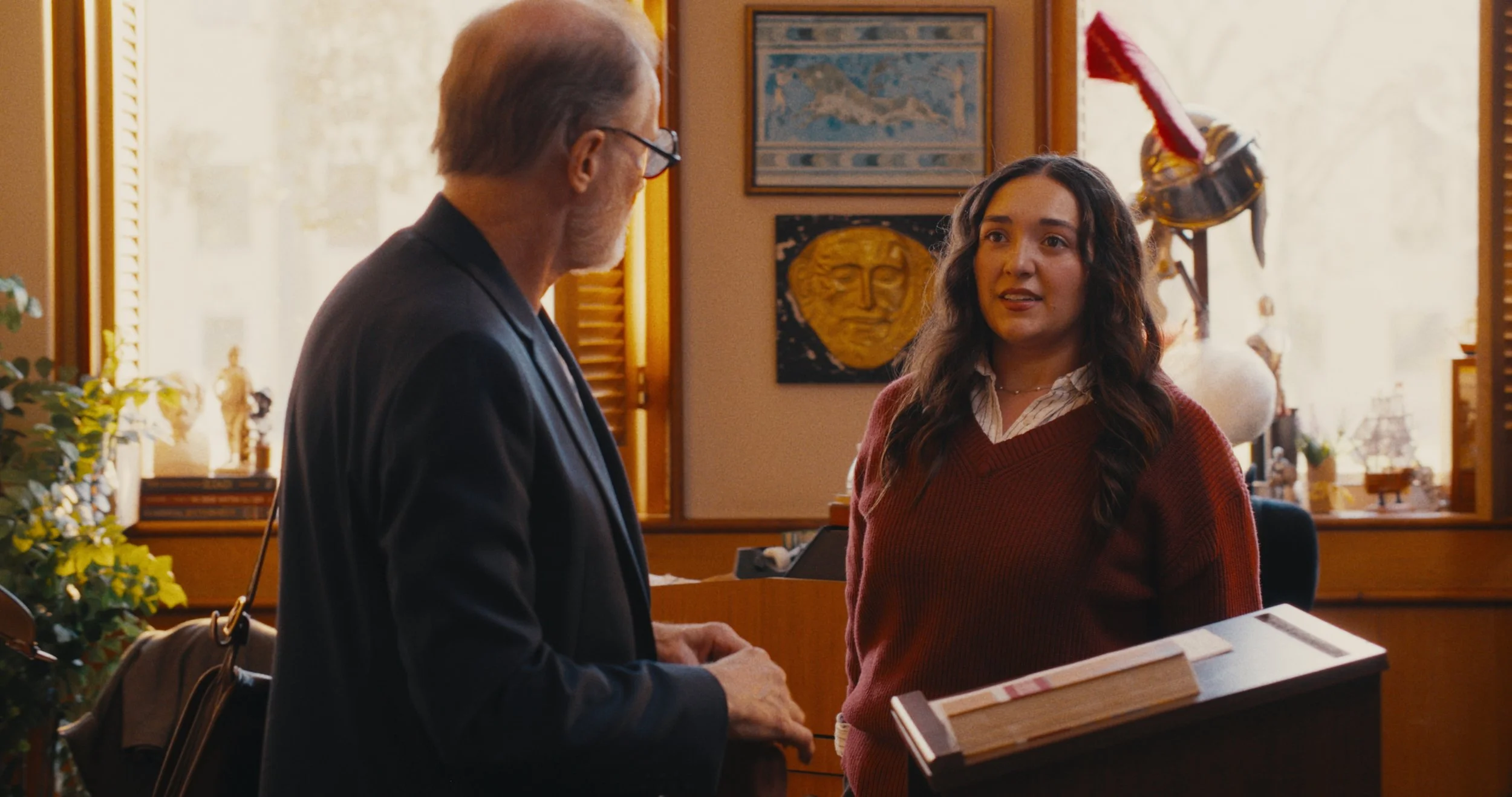 An older man with glasses dressed in a black blazer and a young woman in a red sweater standing at a reception desk and talking.