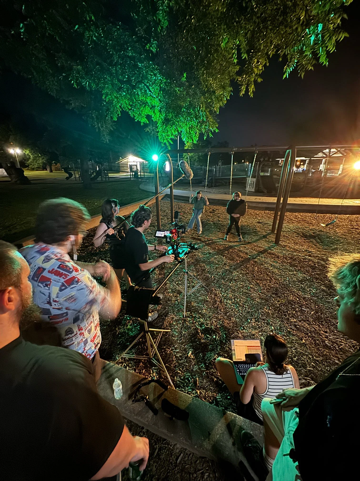 Nighttime scene in a park with children on swings, people sitting on a bench, and a film crew filming the scene with cameras and lighting equipment under a large tree.