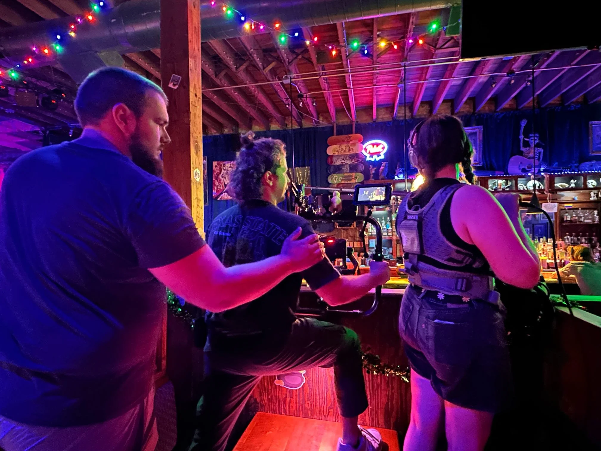 People inside a bar or music venue with colorful string lights and a stage with a neon sign, some people are working with audio or video equipment, and others are socializing.