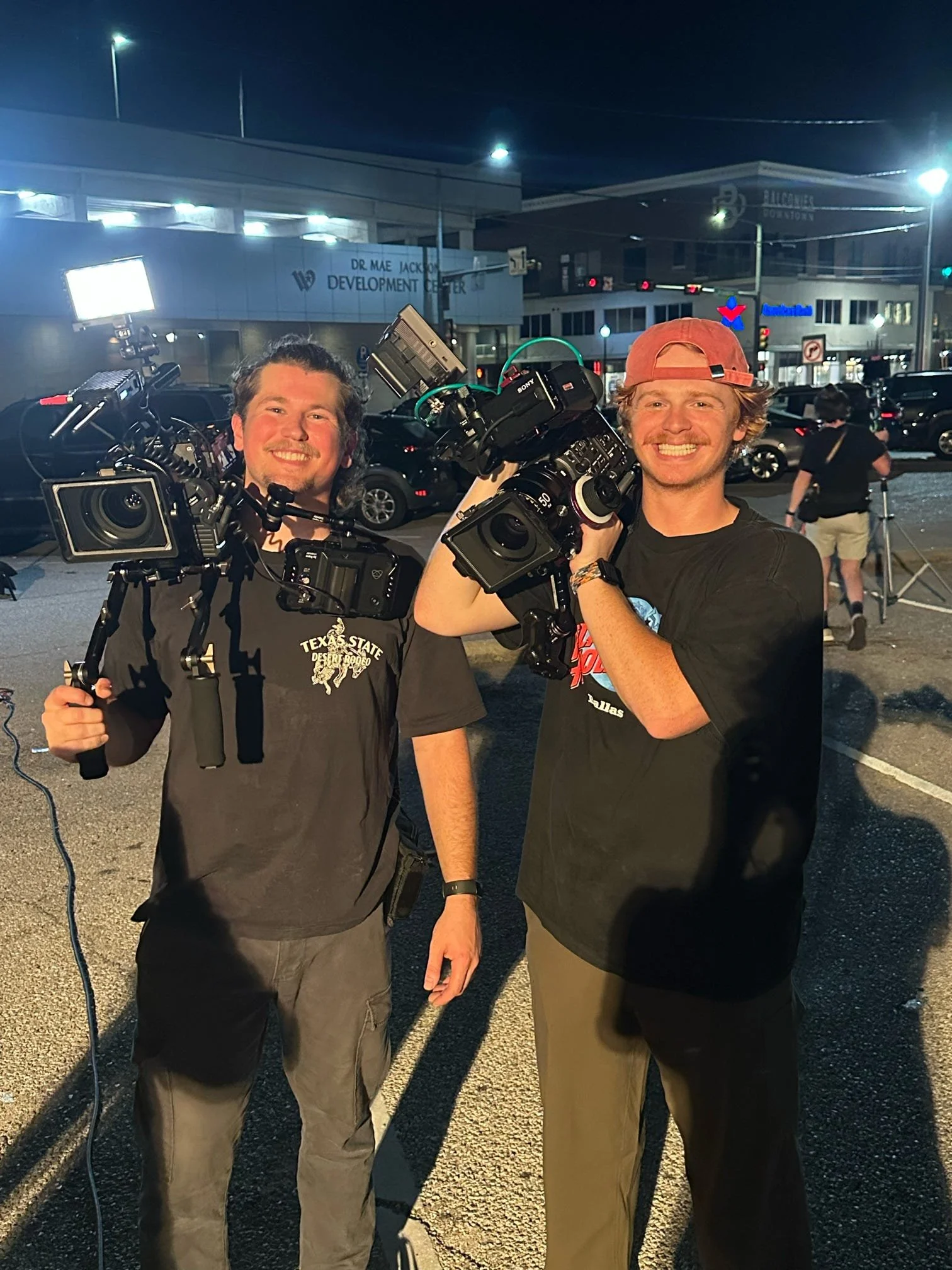 Two men smiling and holding professional video cameras on a city street at night, with streetlights and buildings in the background.