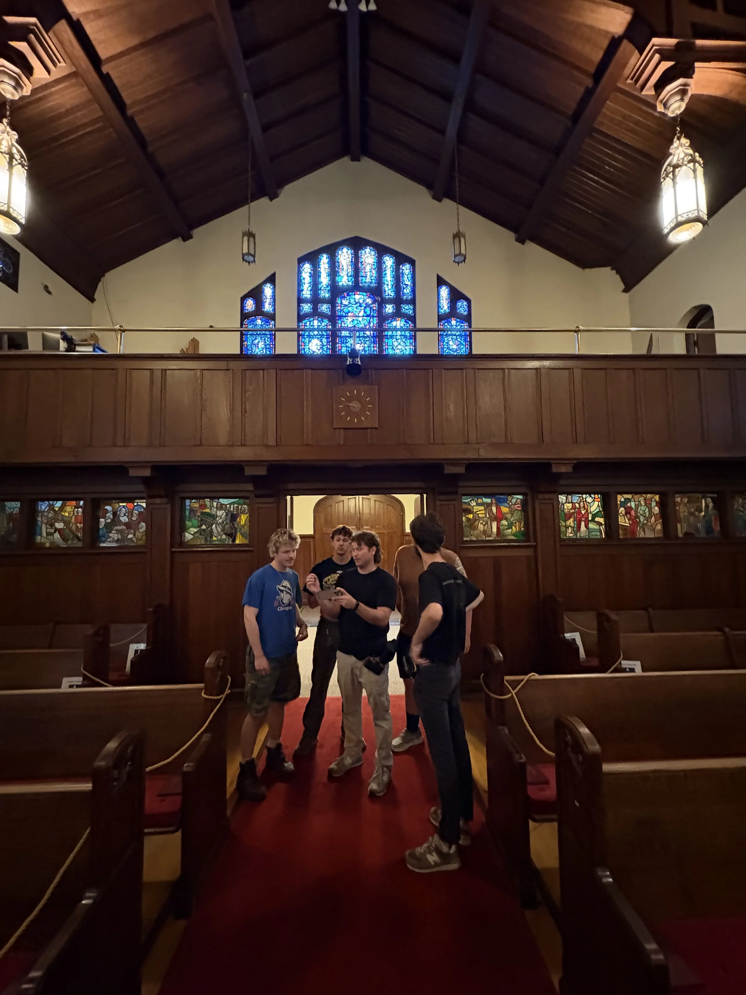 Group of four young men standing and chatting inside a church, with wooden pews, stained glass windows, and a high wooden ceiling.