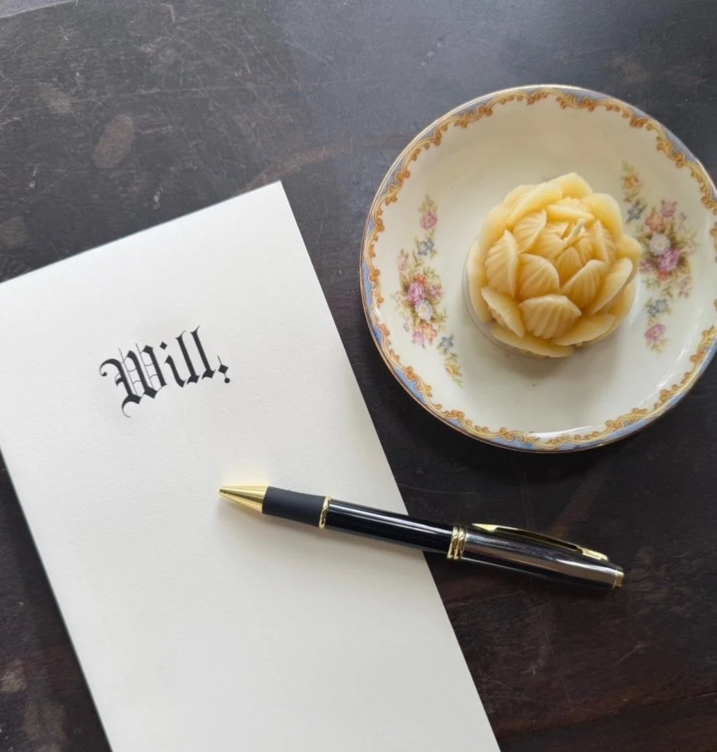 A piece of paper with the word 'Will' written on it, a black and gold pen, and a decorative floral dish holding a wax seal stamp with a cluster of white flowers.