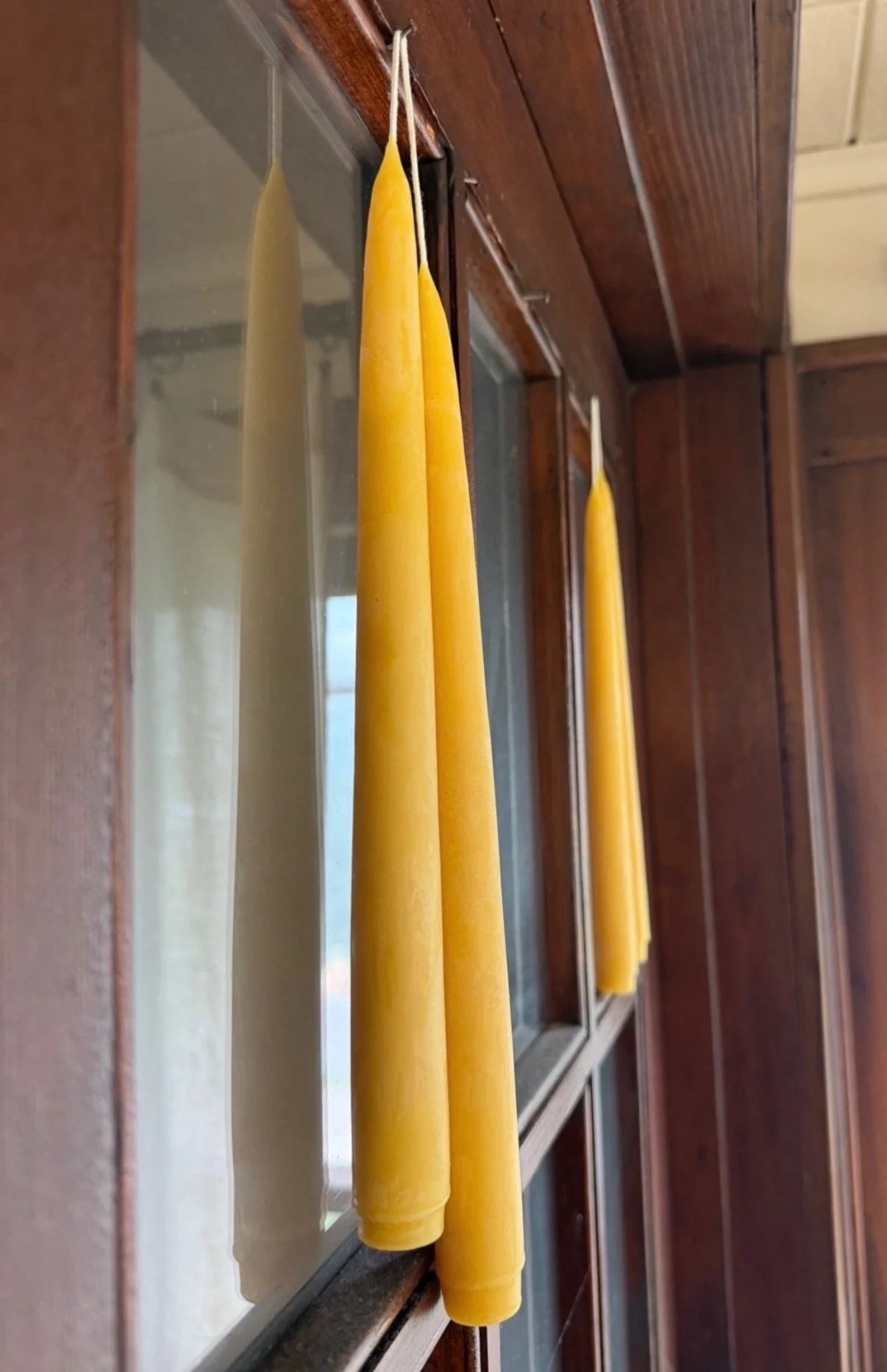 Two gold beeswax candles hanging on hooks inside a wooden window frame.