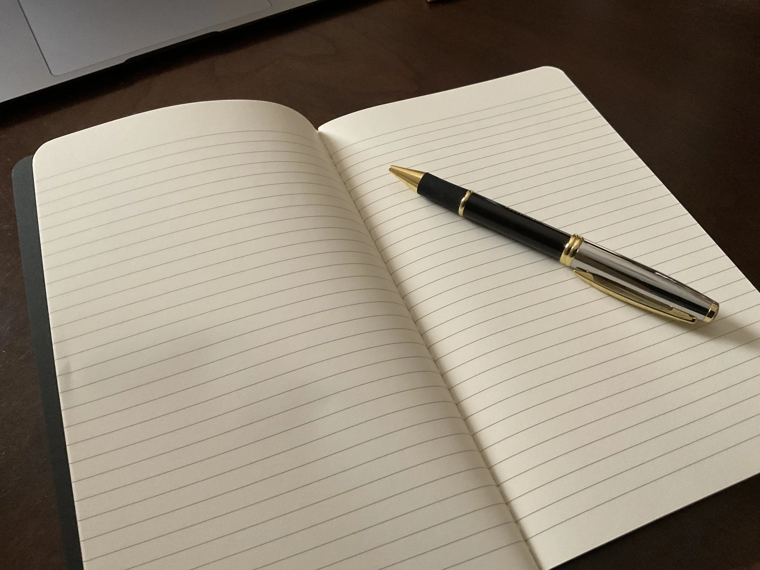 An open blank notebook with lined pages on a dark wooden desk, with a black and gold pen resting on the right page.