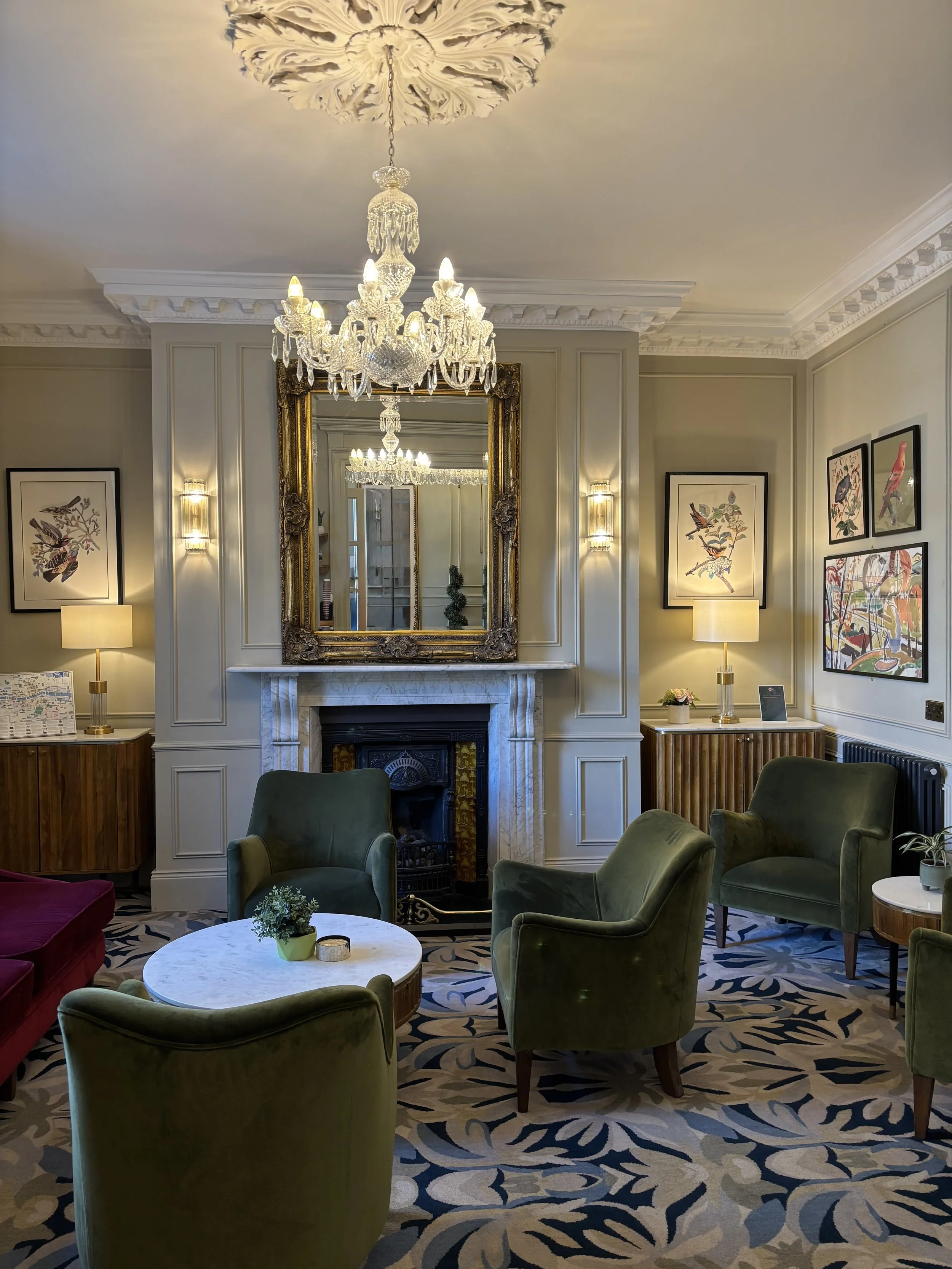A cozy living room with a white marble fireplace, a large ornate gold-framed mirror above it, and a chandelier hanging from the ceiling. The room has green velvet armchairs, a round marble coffee table with a small plant, and framed bird artwork on the walls. The room is warmly lit with table lamps and wall sconces.