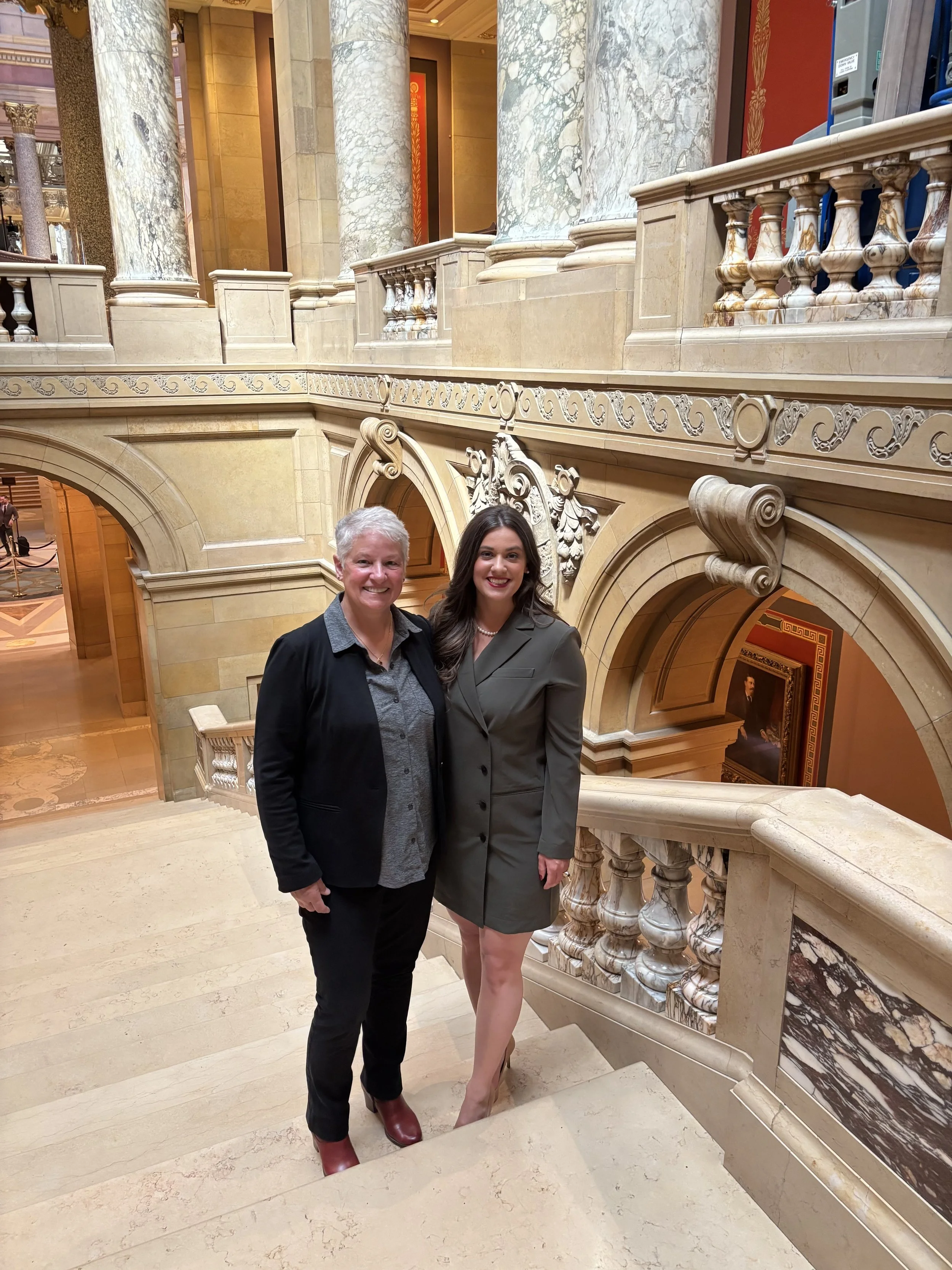 Two women standing on a marble staircase in an ornate interior with marble columns and decorative details.