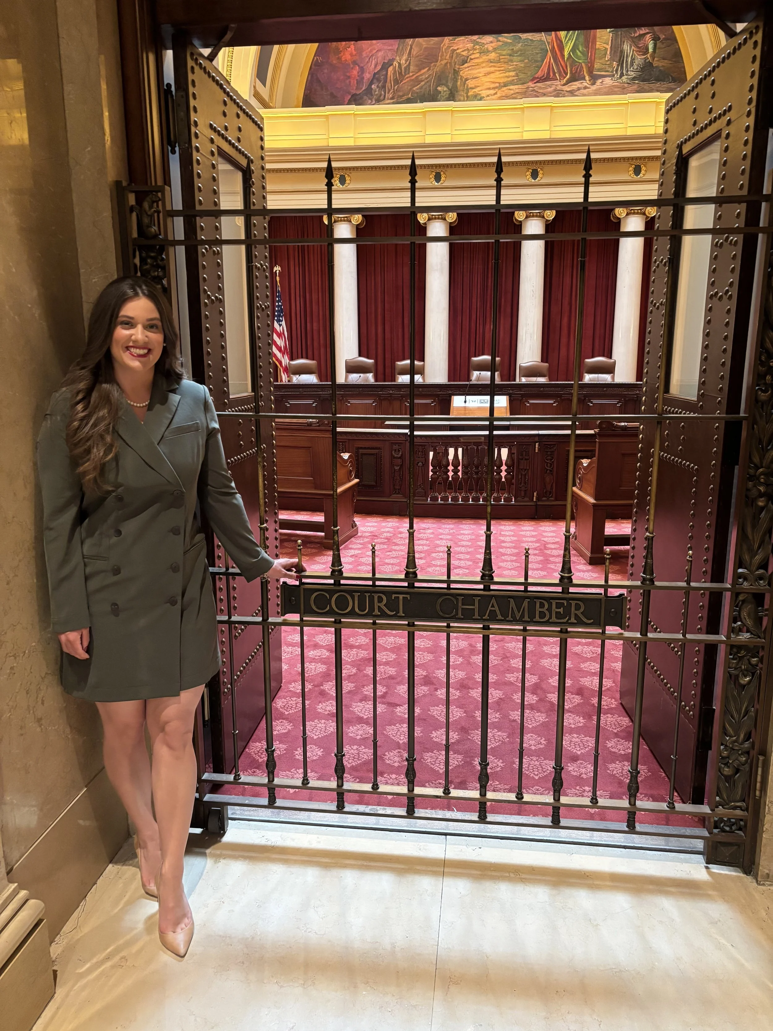 A woman in a gray suit standing beside a gate labeled 'Court Chamber' inside a government building with red carpet and columns in the background.
