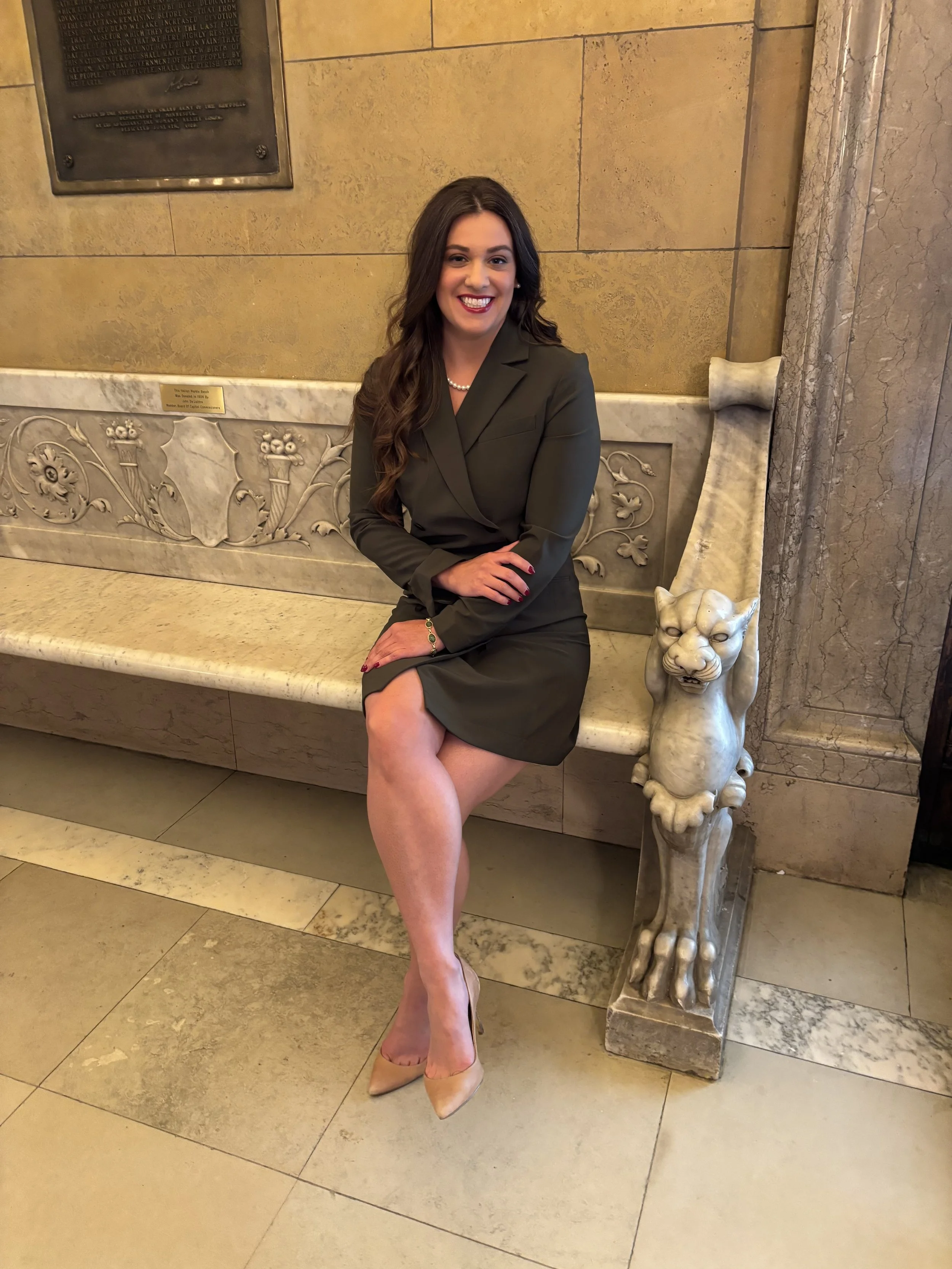 A woman sitting on a marble bench with a lion sculpture at the corner, smiling in a formal suit with beige heels in an indoor setting featuring beige and marble walls.