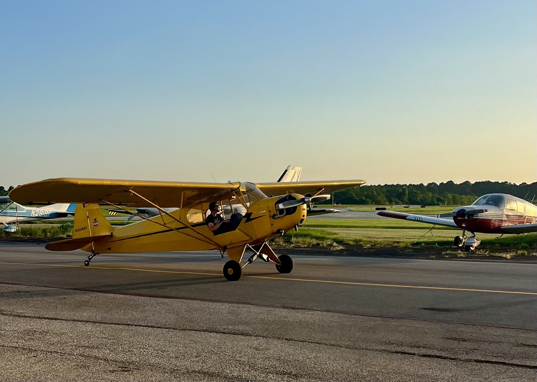A small yellow airplane with a person inside, taxiing on the runway, with a red and white airplane parked in the background on a clear day at an airport.