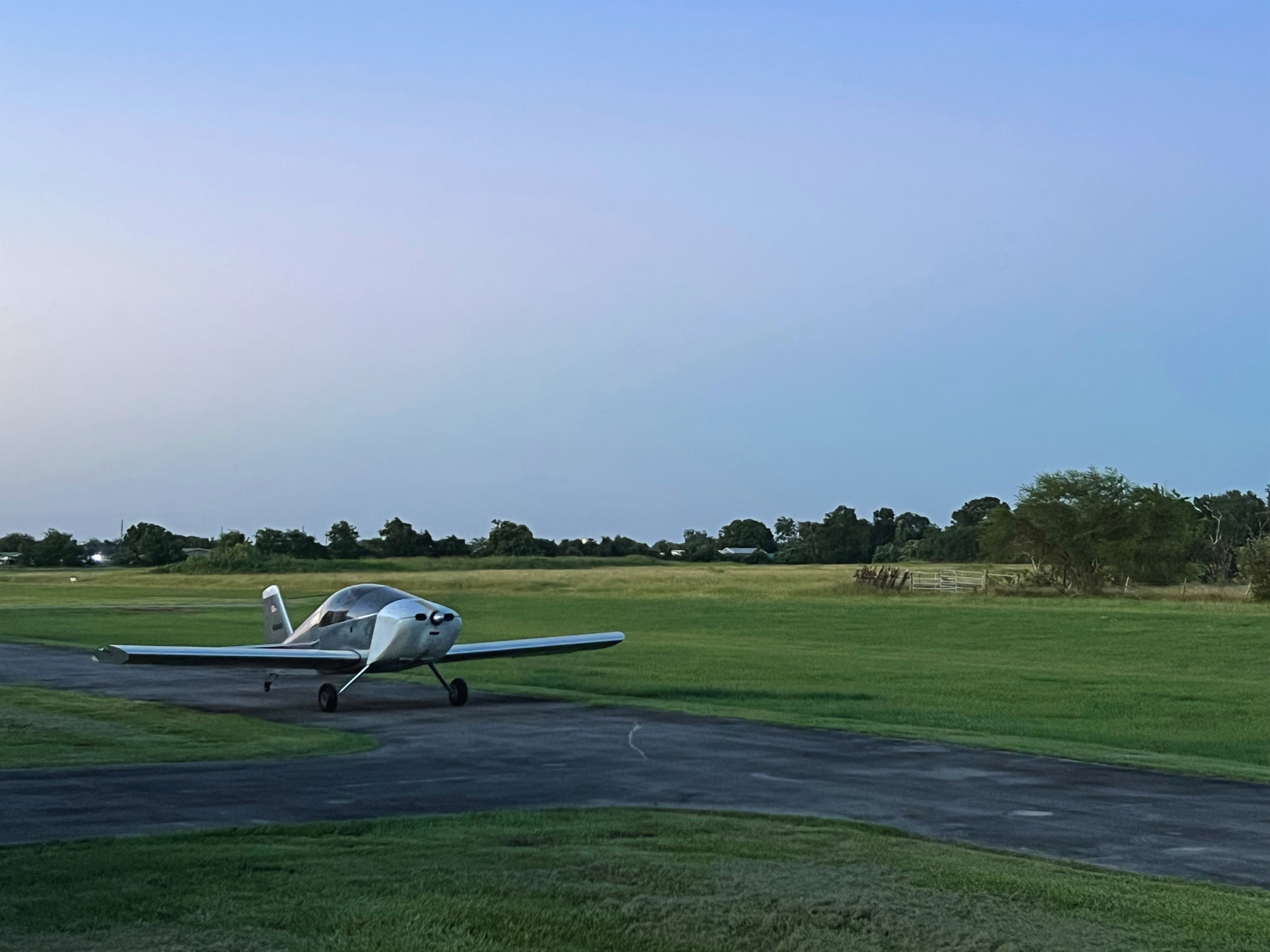 Small white airplane parked on a grassy airfield with trees and buildings in the distance, under a partly cloudy sky.