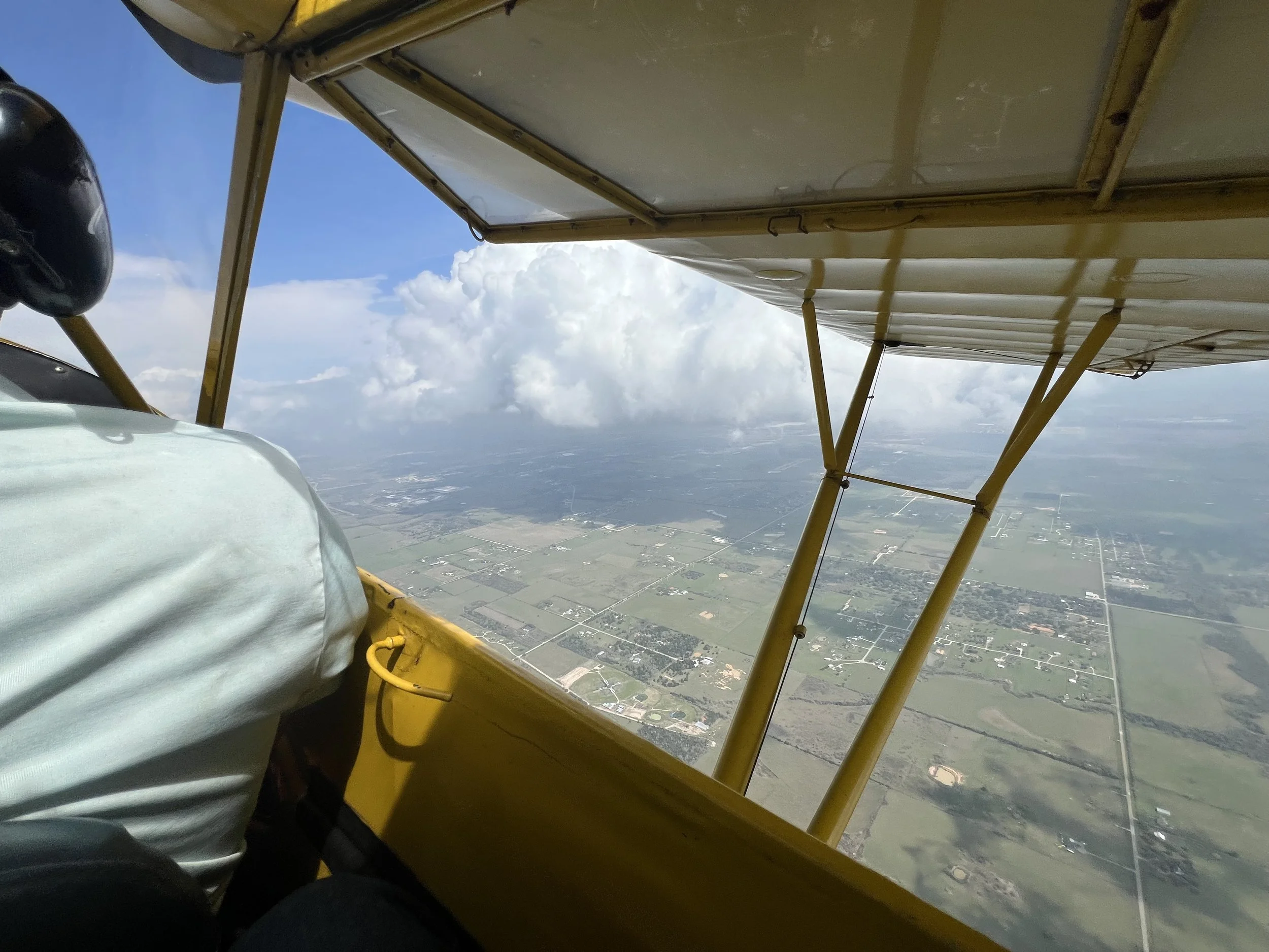 Photo taken from inside a small aircraft during flight shows a view of the aircraft's wing and the landscape below featuring farmland, roads, and scattered structures. The sky is partly cloudy.
