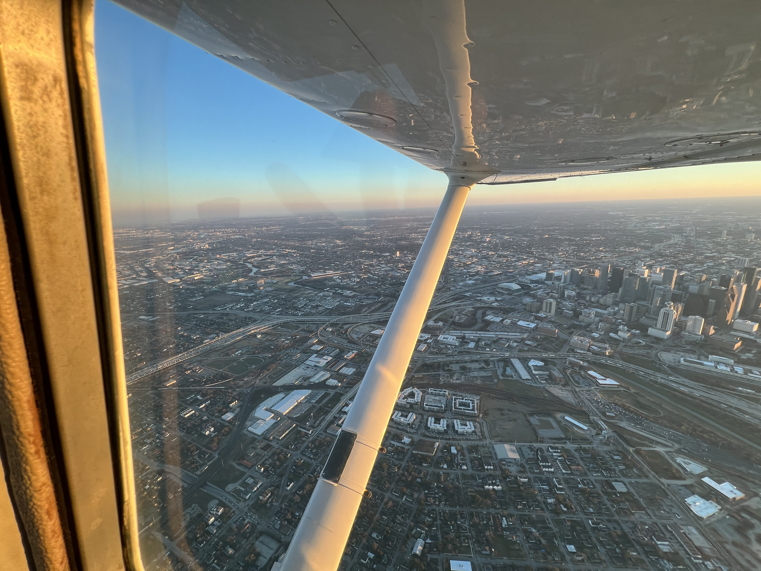 The image shows a view from an airplane flying over a city at sunset. The airplane's wing and support strut are visible, with the cityscape below featuring numerous buildings, roads, and infrastructure.