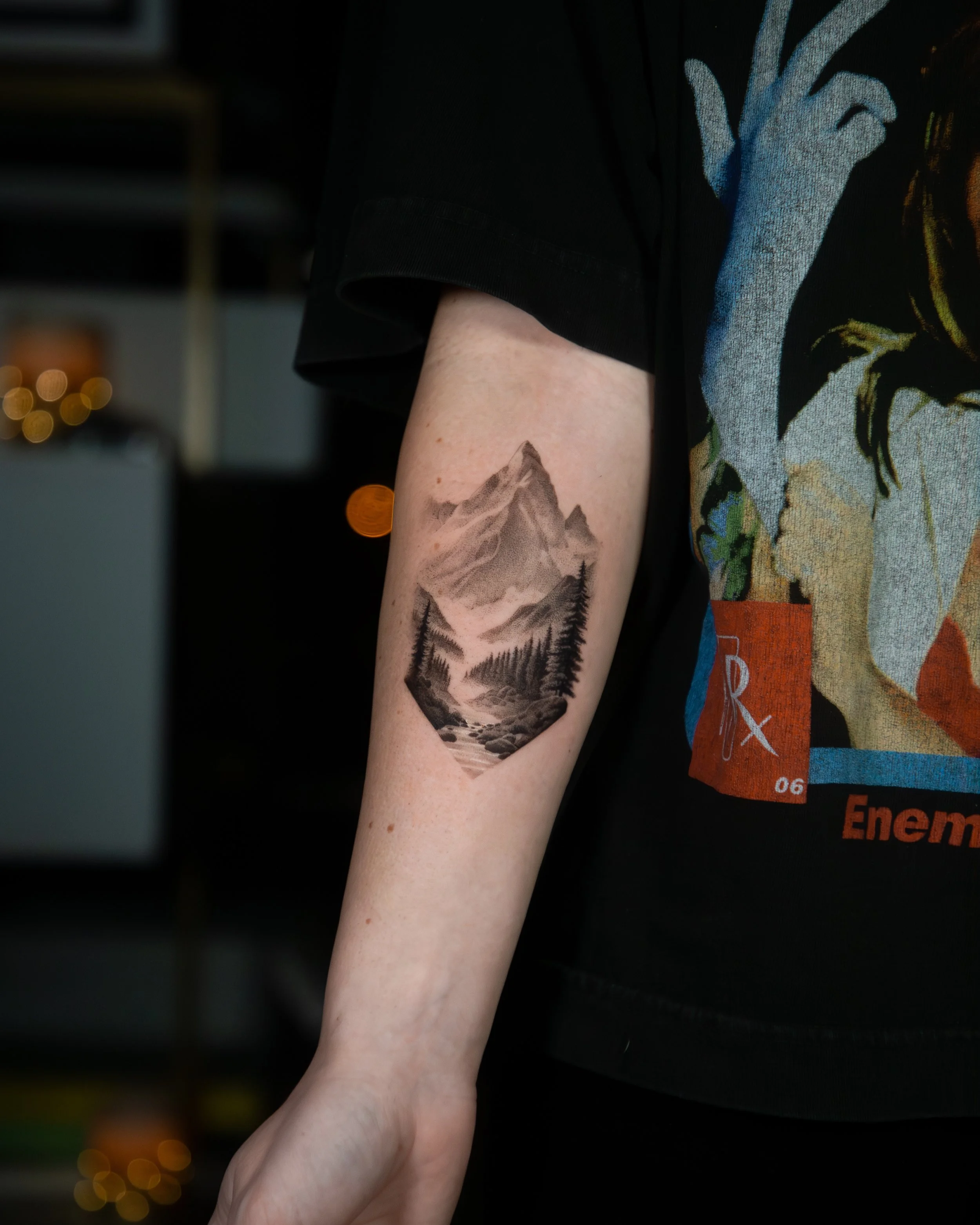 A hyper realistic tattoo done by Ihor (flowerhead.ink) on a person's forearm. Featuring a detailed mountain range, river and pine trees.