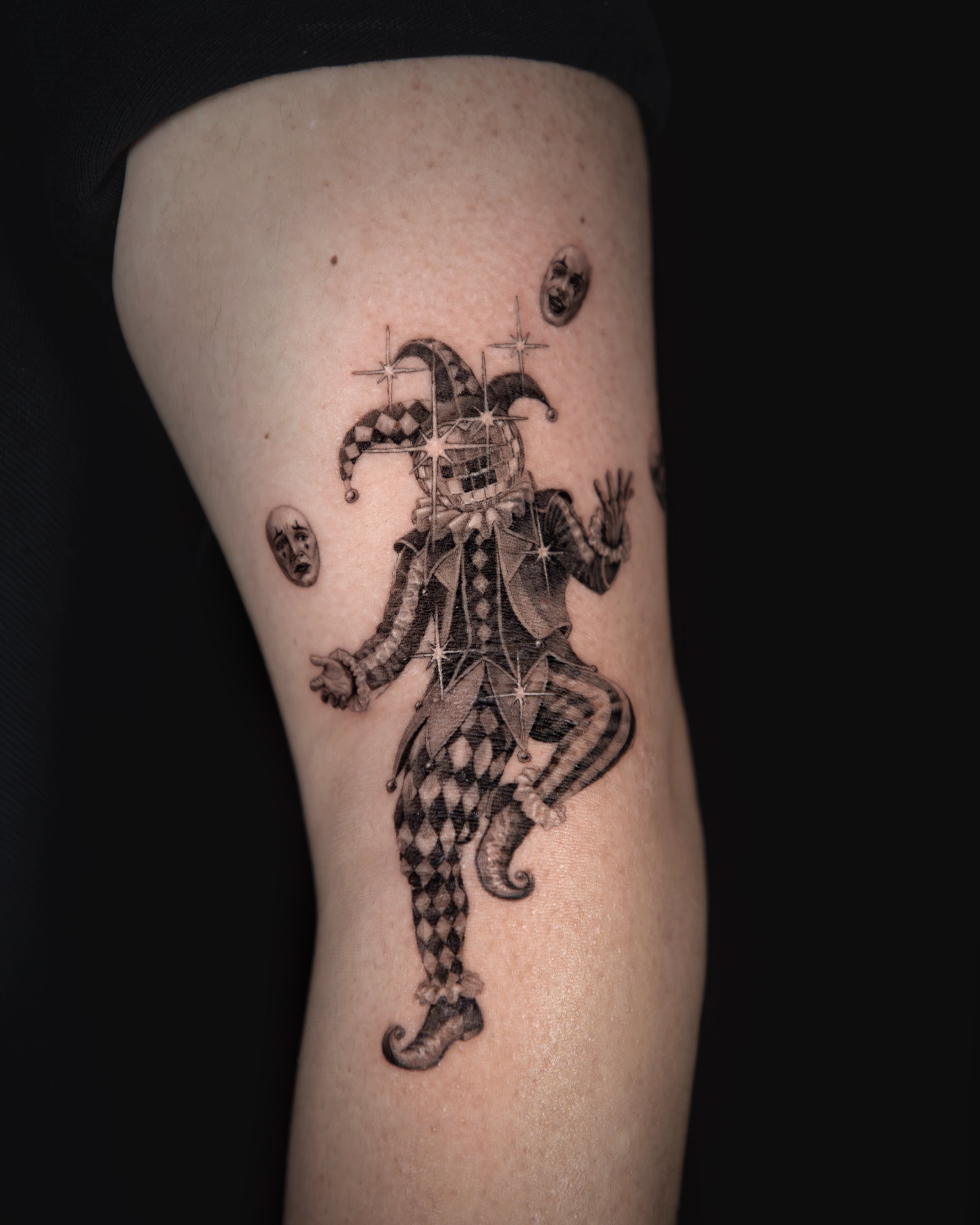 A hyper realistic tattoo done by Ihor (flowerhead.ink) on person's elbow. Featuring a jester with a disco ball as a head, joggling with three face masks. 