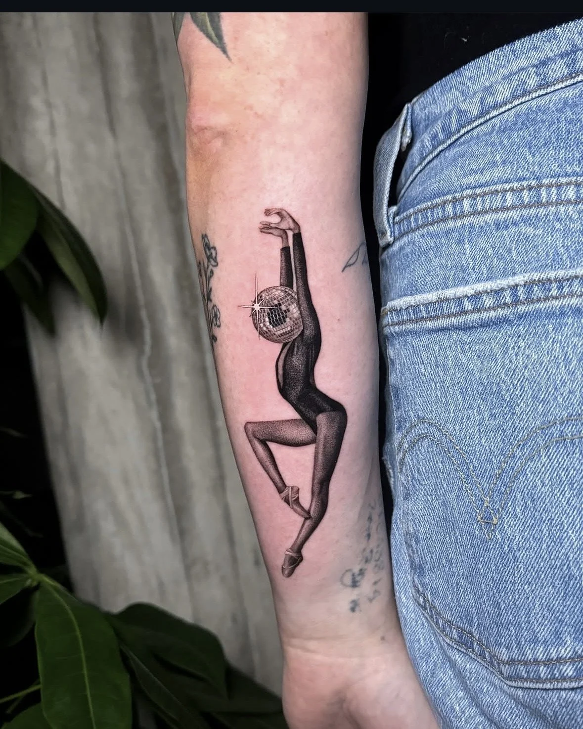 A hyper realistic tattoo done by Ihor (flowerhead.ink) on person's forearm. Featuring a ballerina in fishnet stockings and heels with a disco ball head, dancing in a pose with arms raised and bent legs.