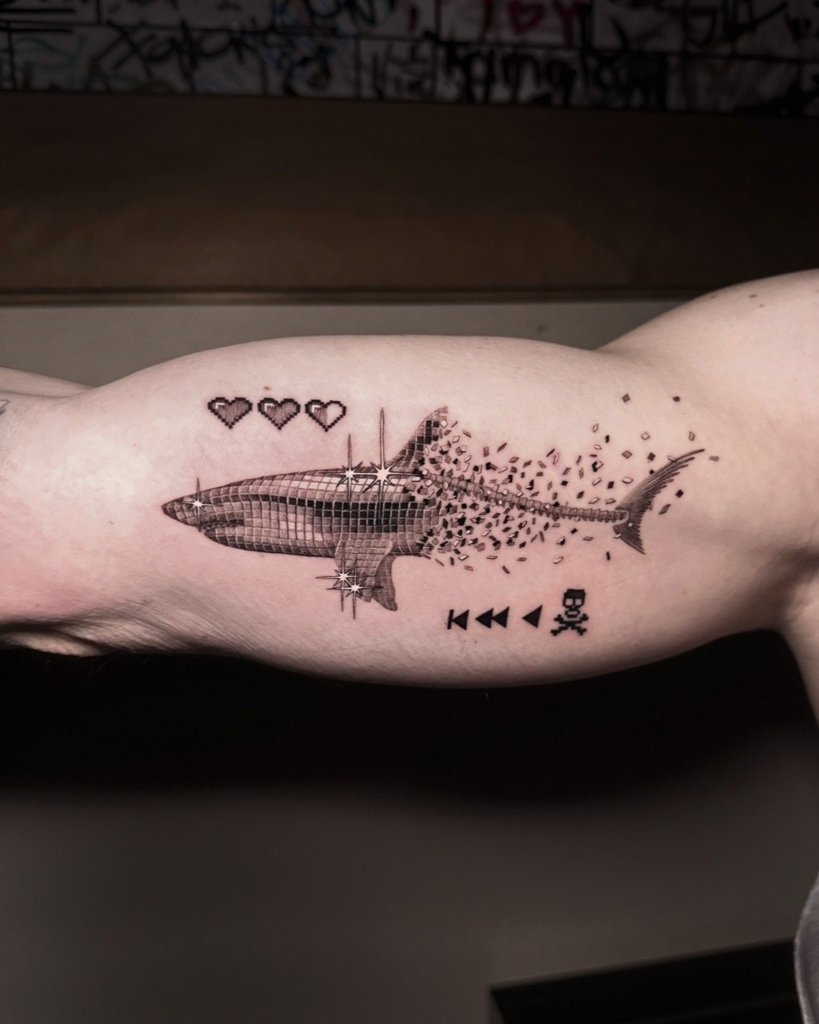 A hyper realistic tattoo done by Ihor (flowerhead.ink) on person's inside biceps. Featuring a shark with a disco ball texture, disintegrating into small squares, with three pixelated hearts above it, and music control symbols below it.