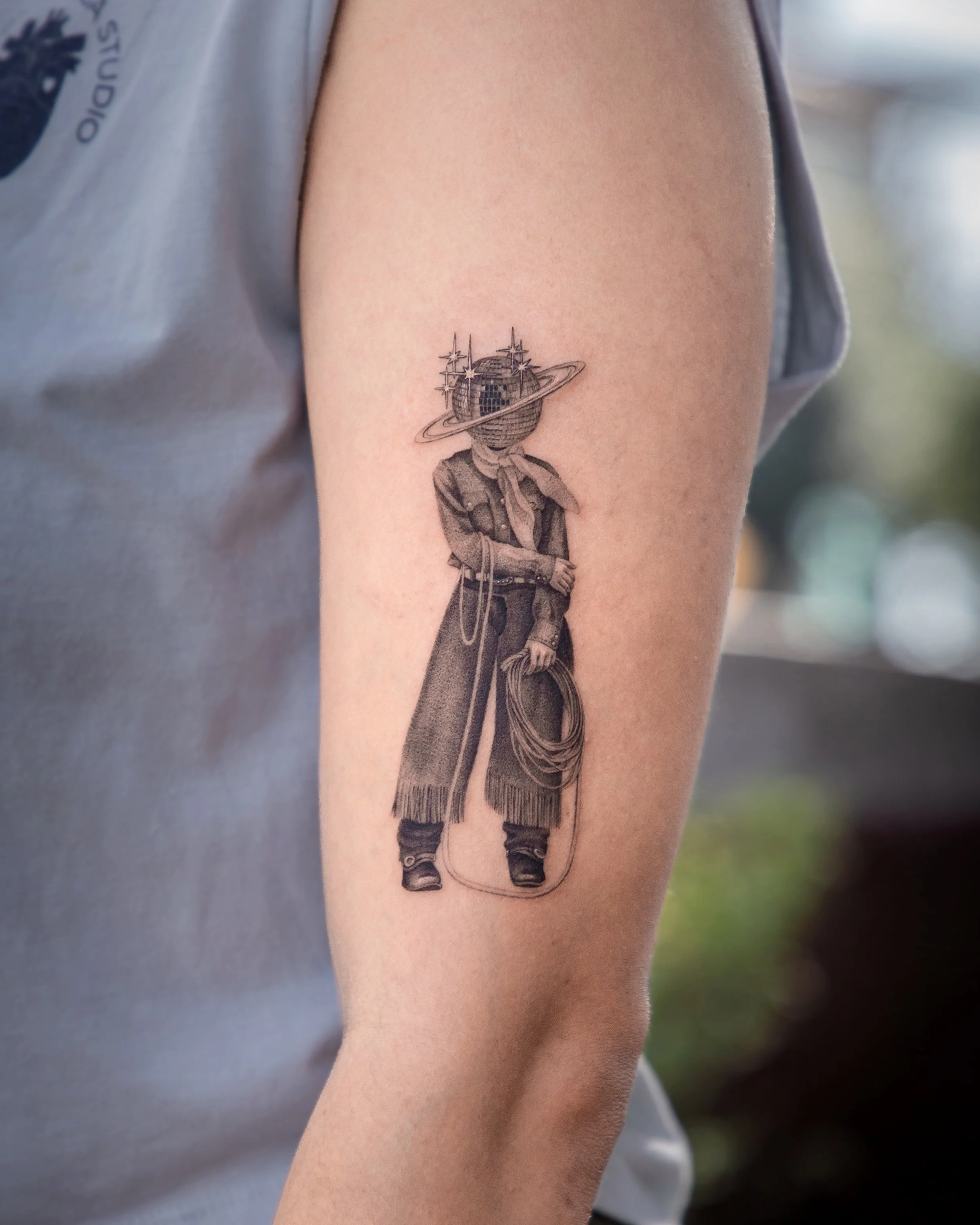 A hyper realistic tattoo done by Ihor (flowerhead.ink) on a person's shoulder. Featuring a person wearing a bandana and old-fashioned cowgirl clothing, a disco ball for a head and a Saturn ring around it.
