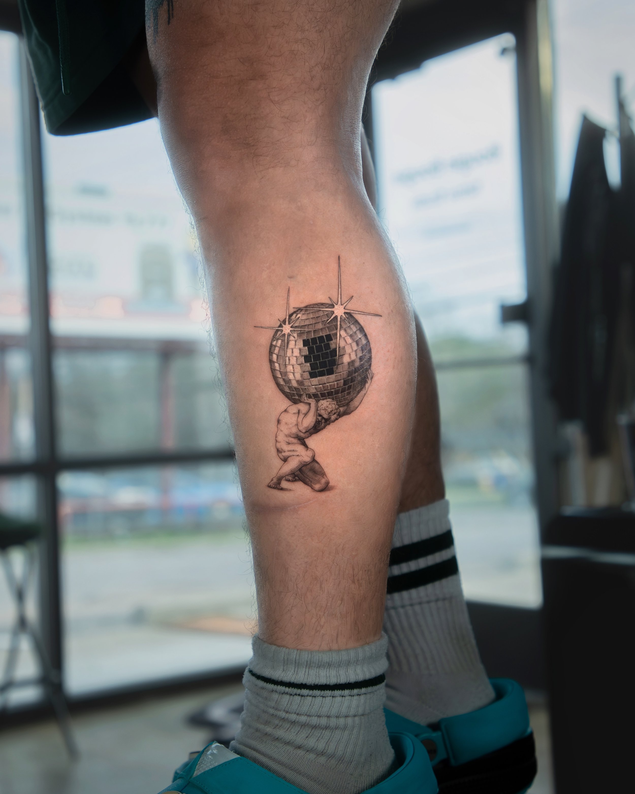 A hyper realistic tattoo done by Ihor (flowerhead.ink) on person's calf. Featuring a traditional sketch of Atlas holding the world, with a disco ball as a planet. 