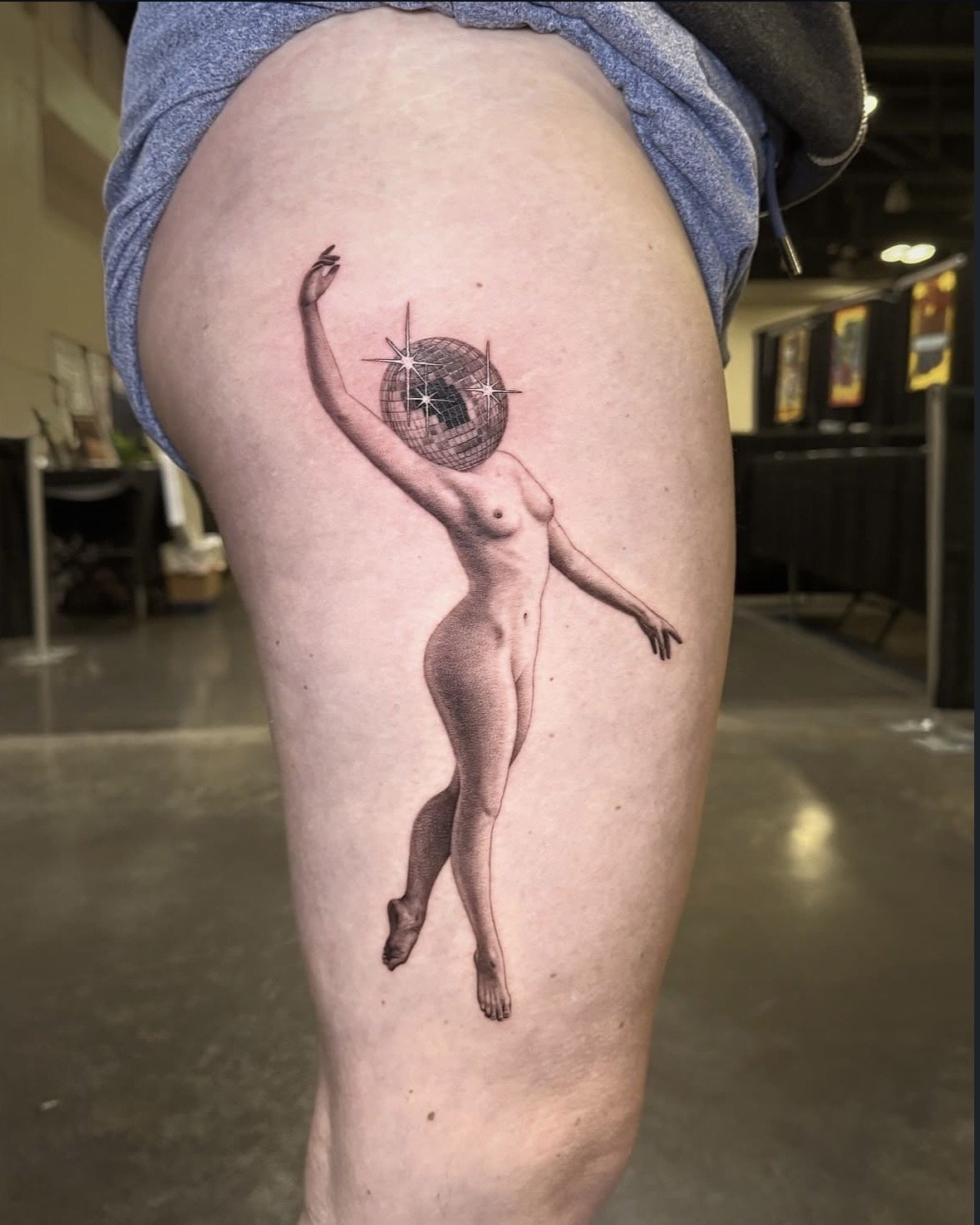 A hyper realistic tattoo done by Ihor (flowerhead.ink) on person's thigh. Featuring a nude woman with a disco ball head and sparkles, standing with arms raised.
