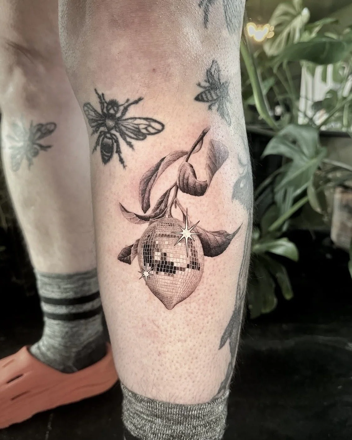 A hyper realistic custom tattoo done by Ihor (flowerhead.ink) on person's calf, below the knee. Featuring an original disco ball textured lemon on a branch with leaves. Dynamic pose. 