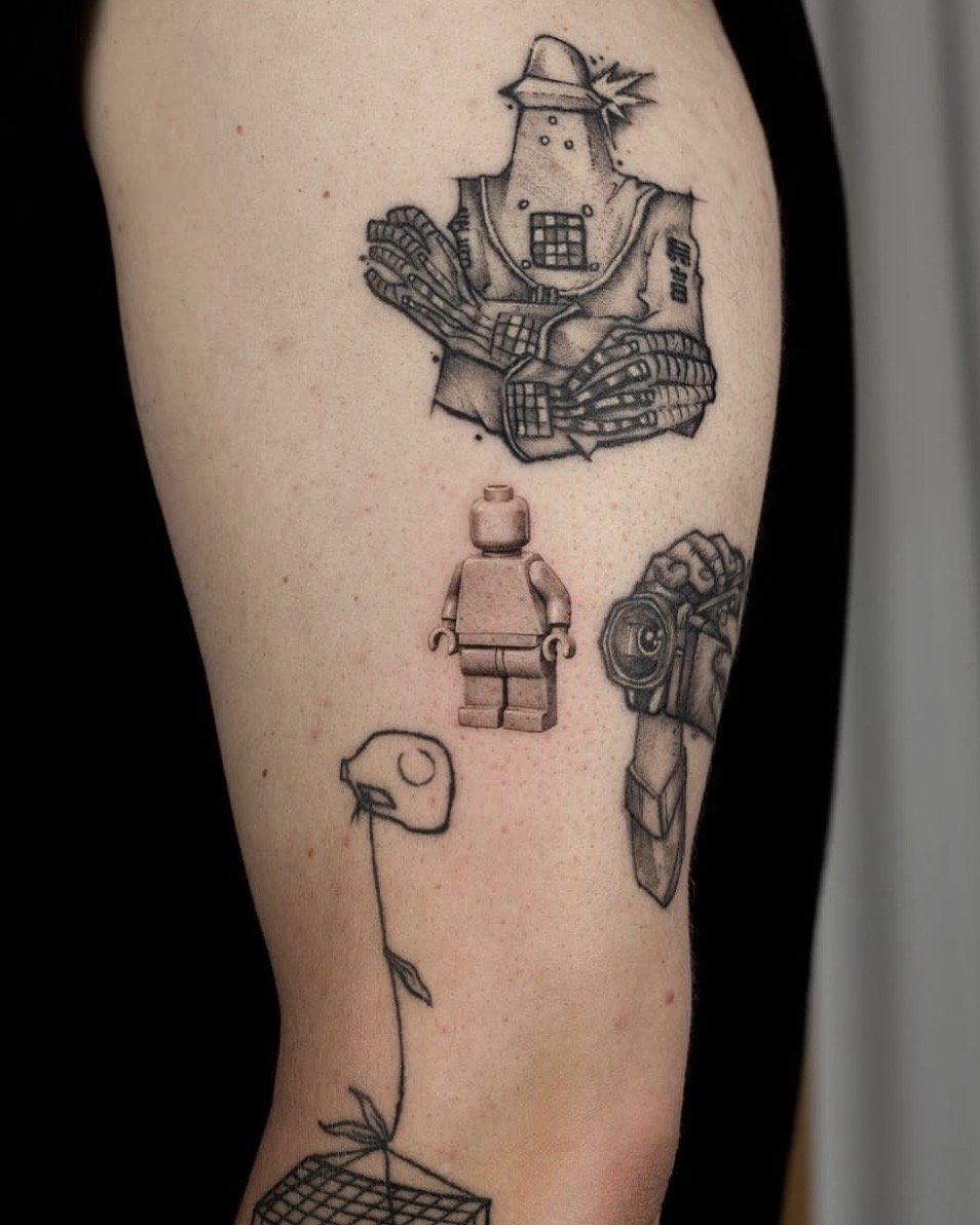 Tattoo by Ihor (flowerhead.ink) on a person's shoulder, featuring a LEGO mini-figure in hyper realism style, that looks objectively better then the other tattoos around it.