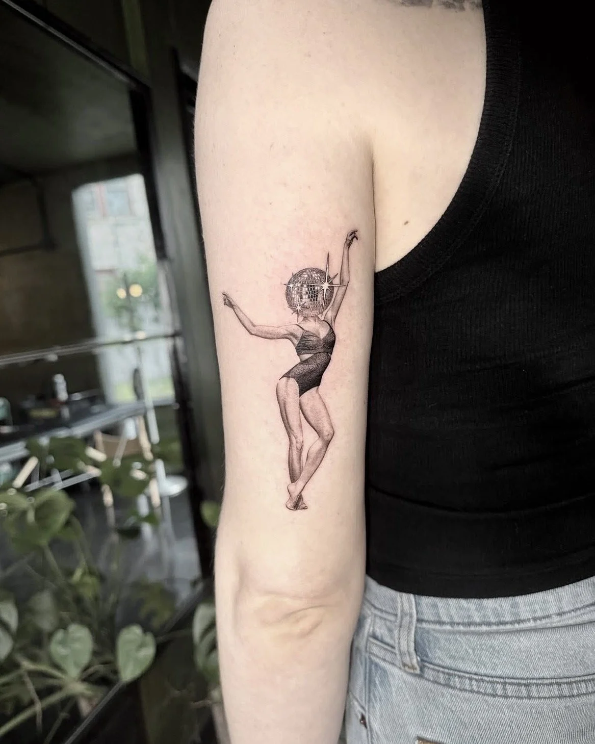 A hyper realistic tattoo done by Ihor (flowerhead.ink) on person's upper arm. Featuring a dancing woman with a disco ball for a head, wearing a swimsuit.