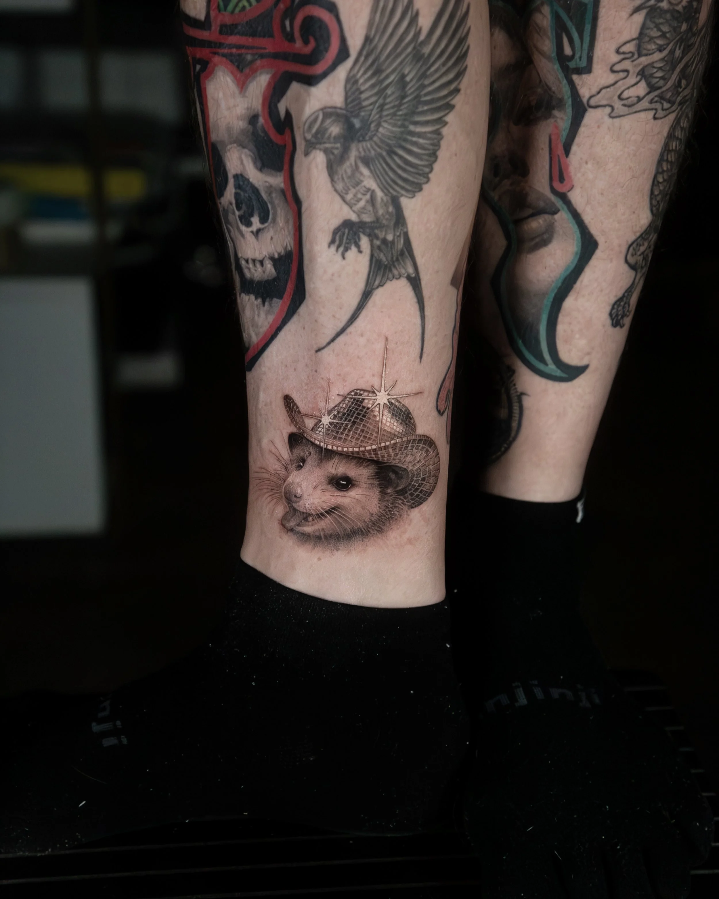 A hyper realistic tattoo done by Ihor (flowerhead.ink) on person's ankle. Featuring a portrait of a happy opossum wearing a disco ball hat. 