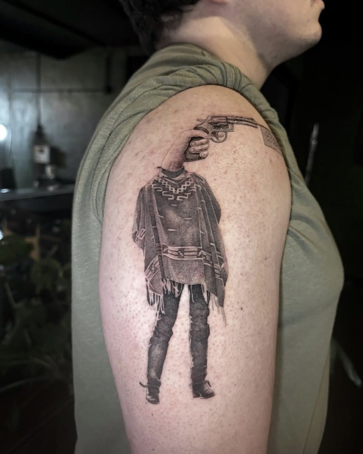 A hyper realistic tattoo done by Ihor (flowerhead.ink) on person's forearm. Featuring a cowboy with a hand holding a gun as his head. Wearing a poncho and boots. The revolver has a "BANG!" sticker coming out of it. 