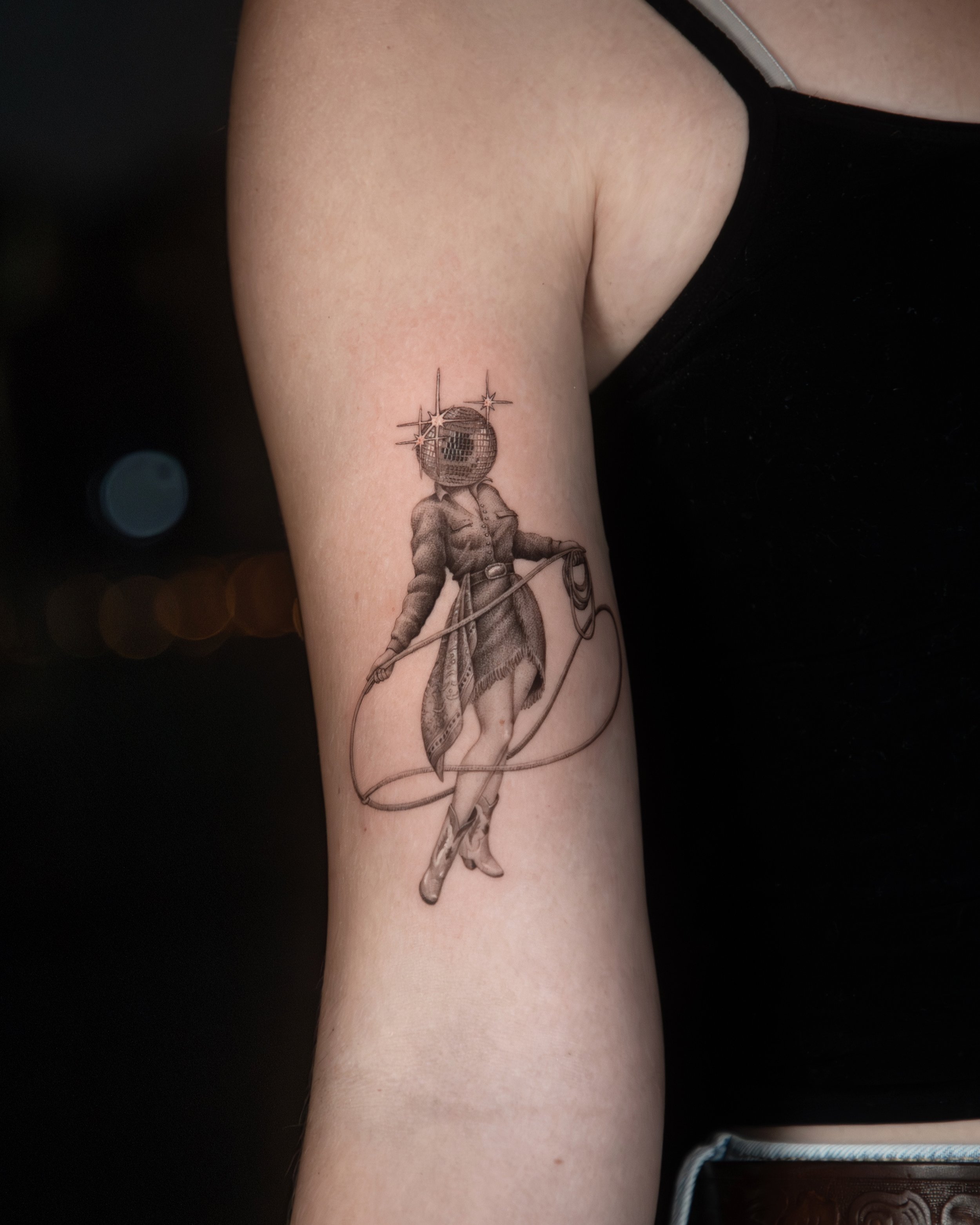 A hyper realistic tattoo done by Ihor (flowerhead.ink) on person's upper arm. Featuring a cowgirl with a shiny disco ball as a head, wearing a vintage dress and holding a laso.