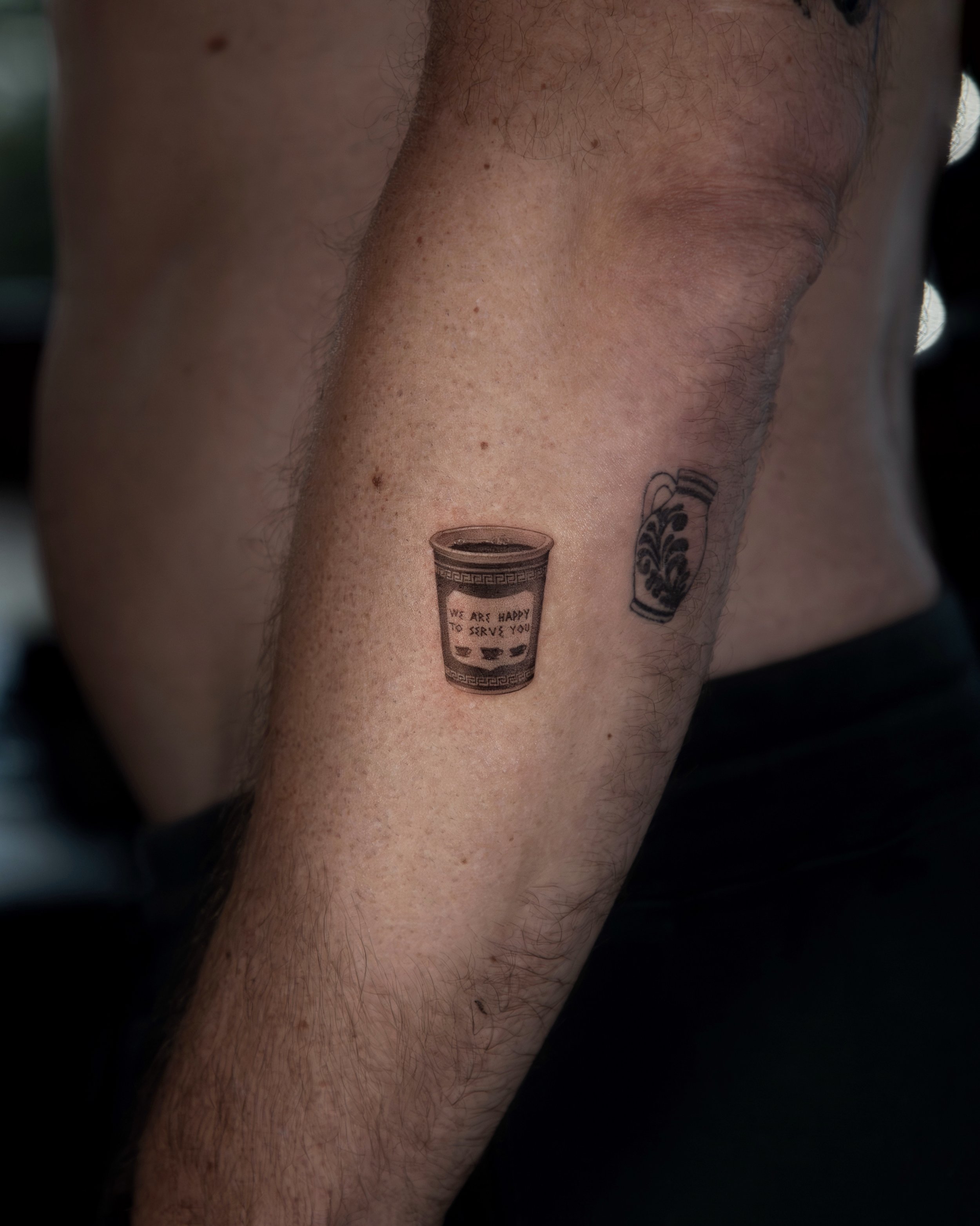 A hyper realistic tattoo done by Ihor (flowerhead.ink) on a person's forearm. Featuring a close-up of a traditional NYC coffee cup with the message 'We are happy to serve you". 