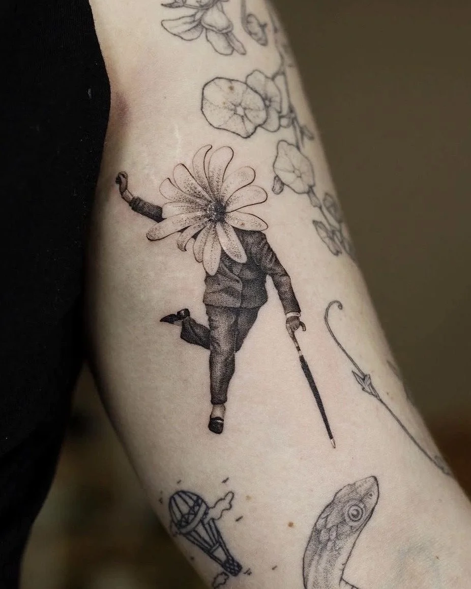 A hyper realistic tattoo done by Ihor (flowerhead.ink) on a person's front of a biceps. Featuring a person with a large flower as a head (flowerhead), dancing and holding an umbrella. 