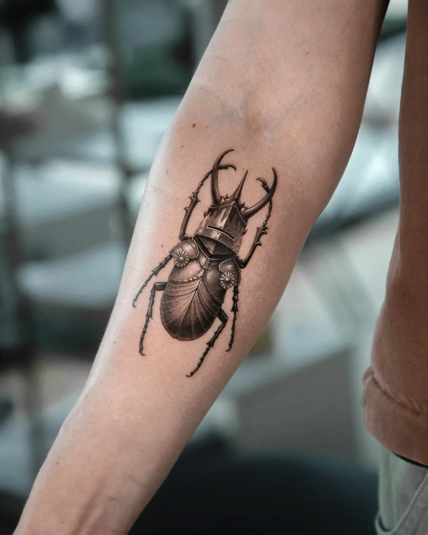 A custom tattoo by Ihor (flowerhead.ink) on a person's forearm, featuring a beetle with shiny medieval armor.