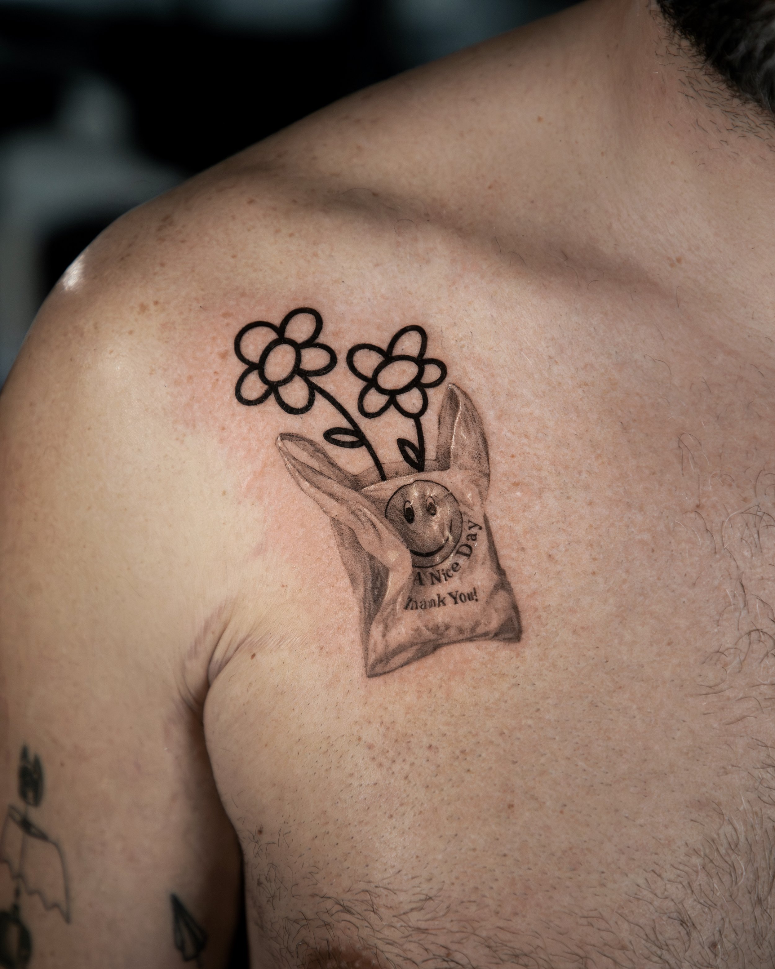 A custom tattoo done by Ihor (flowerhead.ink) on a person's chest. Featuring a traditional NYC shopping bag with a smiling face and a bouquet of two flowers coming out of it. Hyper realistic style with a combination of simple and thick line work. 