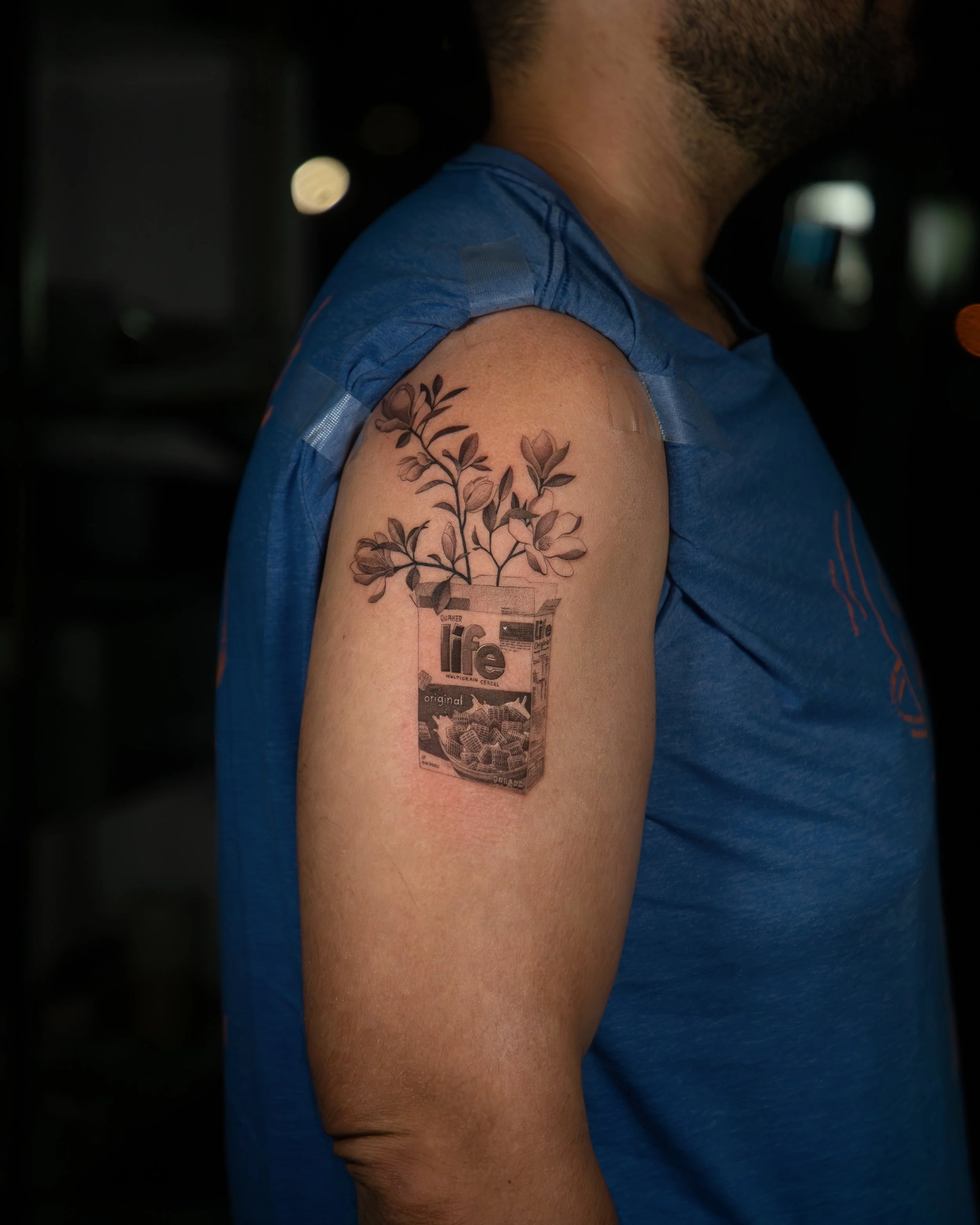 A hyper realistic tattoo done by Ihor (flowerhead.ink) on a person's shoulder. Featuring a Life cereal box with magnolias coming out of it. 