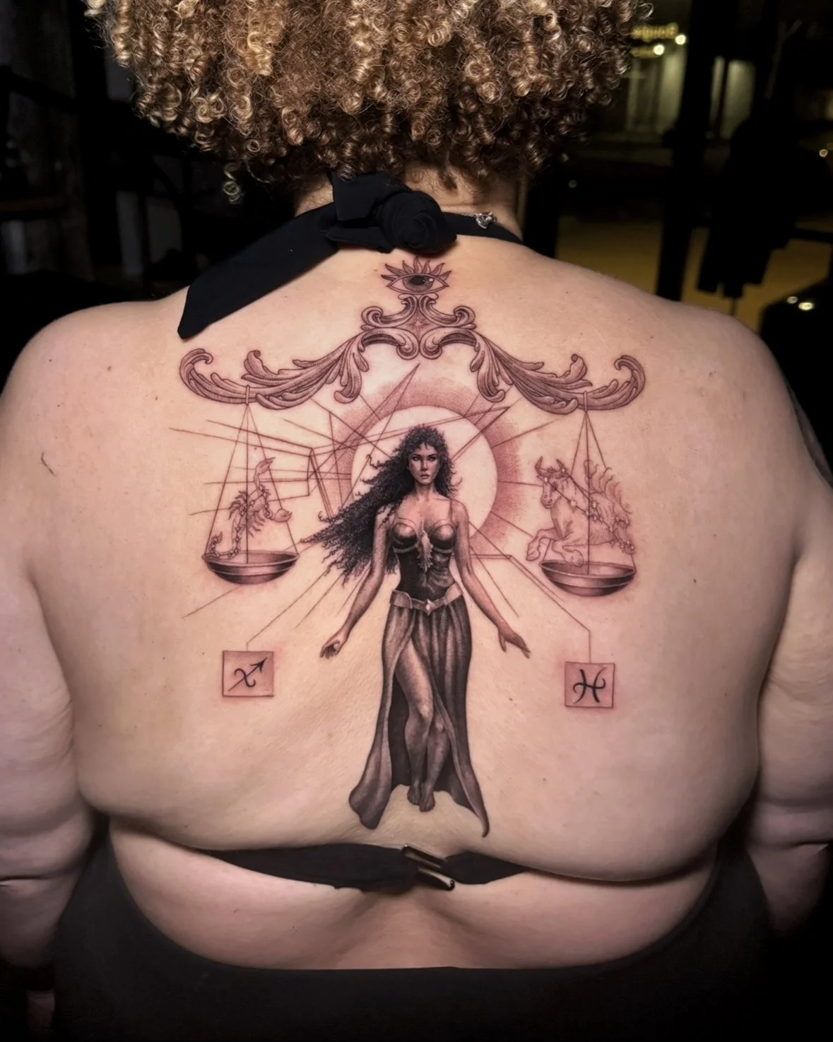 A hyper realistic tattoo done by Ihor (flowerhead.ink) on person's back. Featuring a woman with long curly hair, standing in front of a large balance scale with an eye at the top, flanked by Scorpio and Pisces symbols.